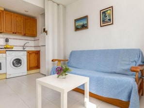 Un espacio habitable compacto con kitchenette, un sofá azul, una mesita con un florero y dos cuadros en la pared.