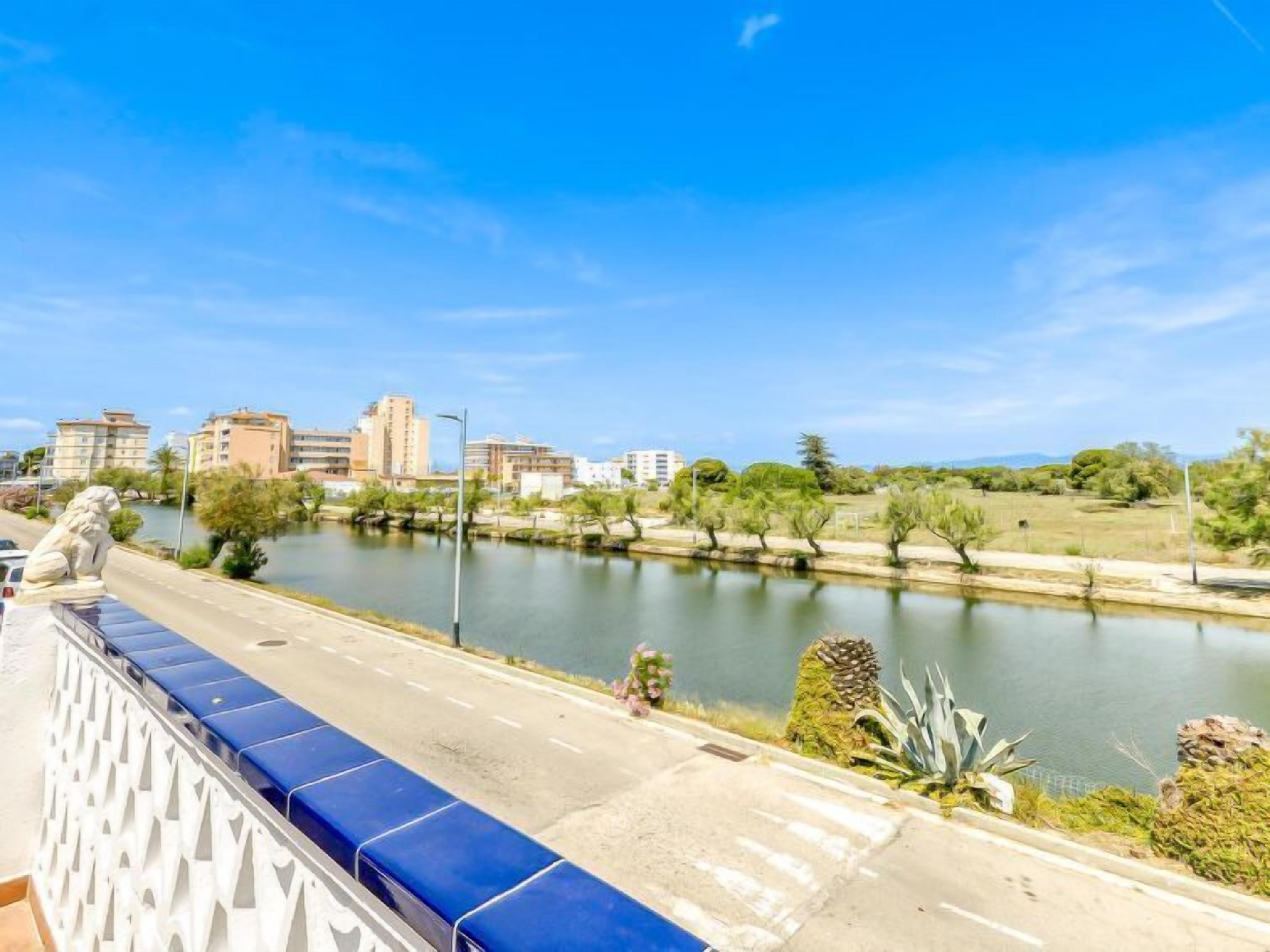 Un río con edificios al fondo, una carretera con una barrera azul y vegetación a lo largo de la ribera.