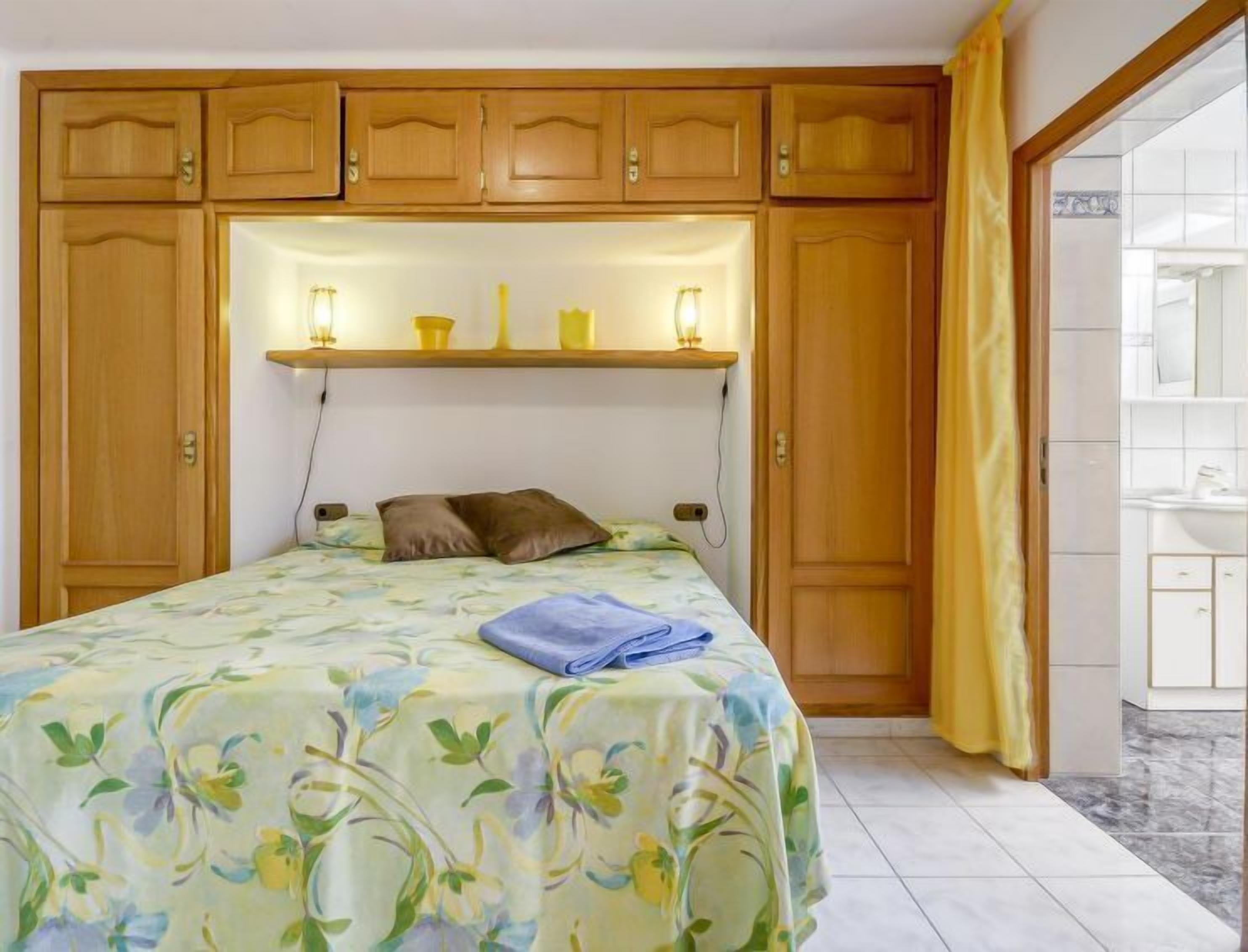 Een slaapkamer met een bed, houten kasten, een verlichte plank en een zichtbare badkamer door een geopende deur.
