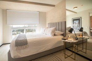 Ein modernes Hotelzimmer mit einem großen Bett, einem Nachttisch mit Lampe, Blick auf die Stadt durch das Fenster und einem gerahmten Bild an der Wand.