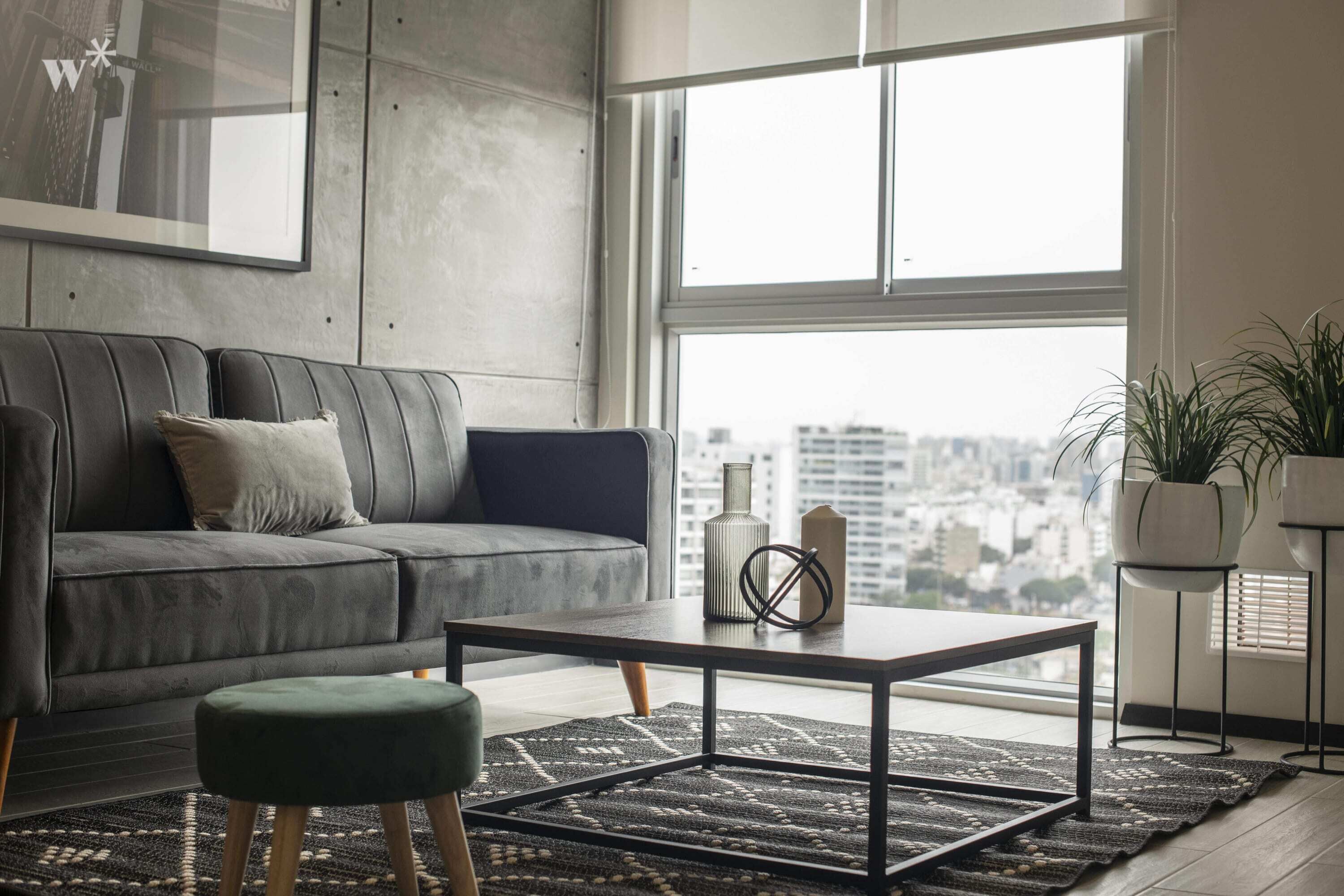 Een moderne woonkamer met een grijze bank, een salontafel en een uitzicht op de stad door grote ramen.