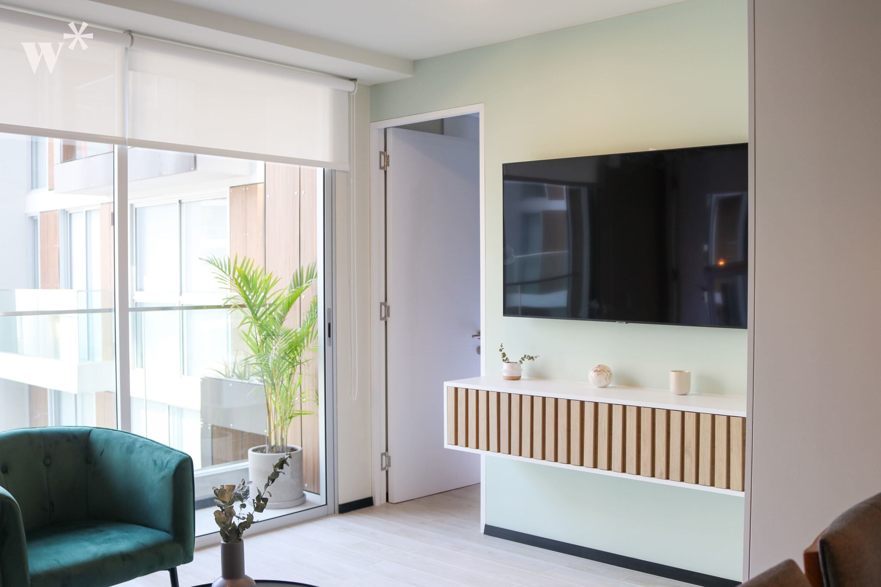 A modern living room with a sliding glass door, a green armchair, a potted plant, a flat-screen TV mounted on a light-colored wall, and a wooden console table.