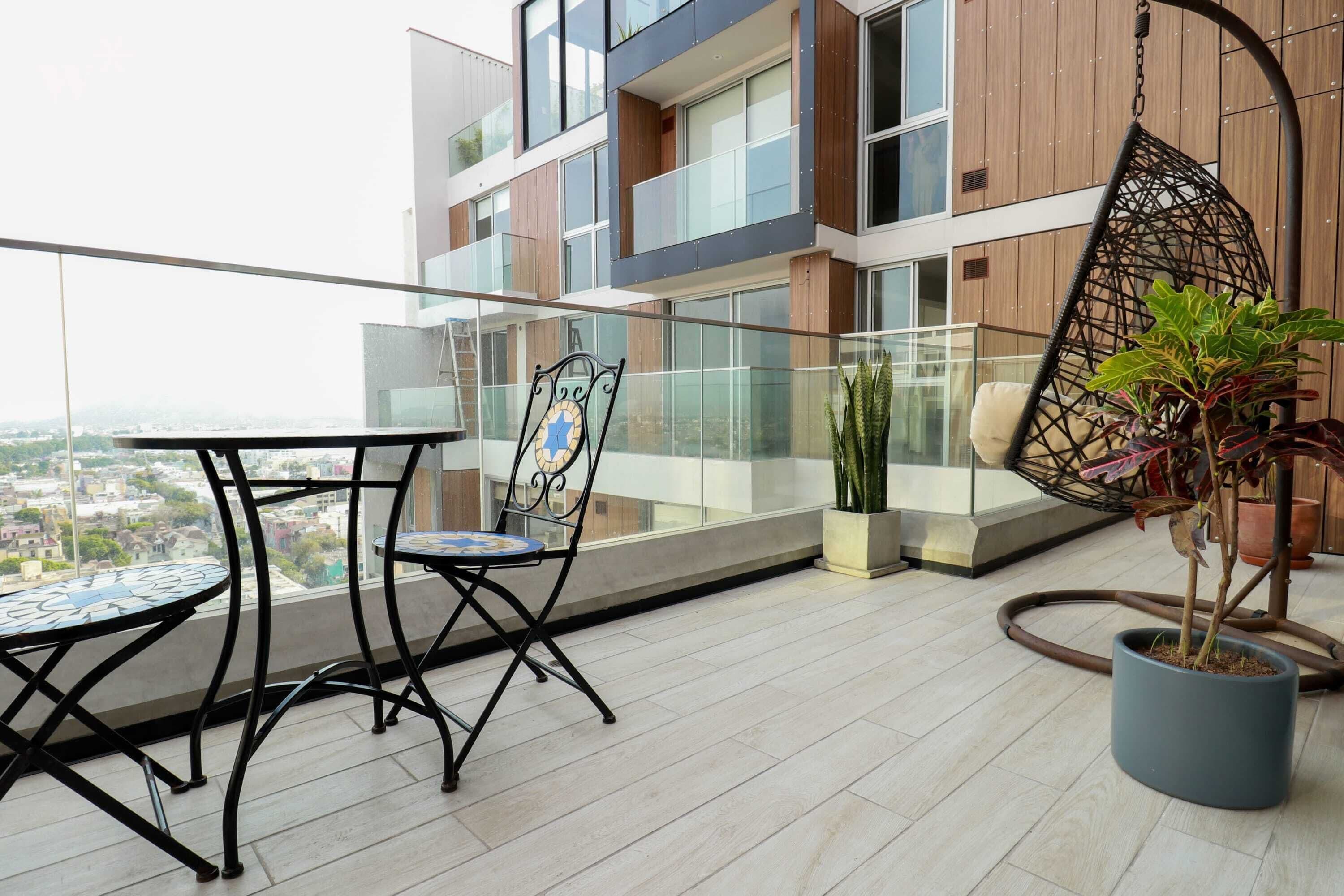 A balcony with a glass railing, a hanging chair, a table with chairs, and potted plants.