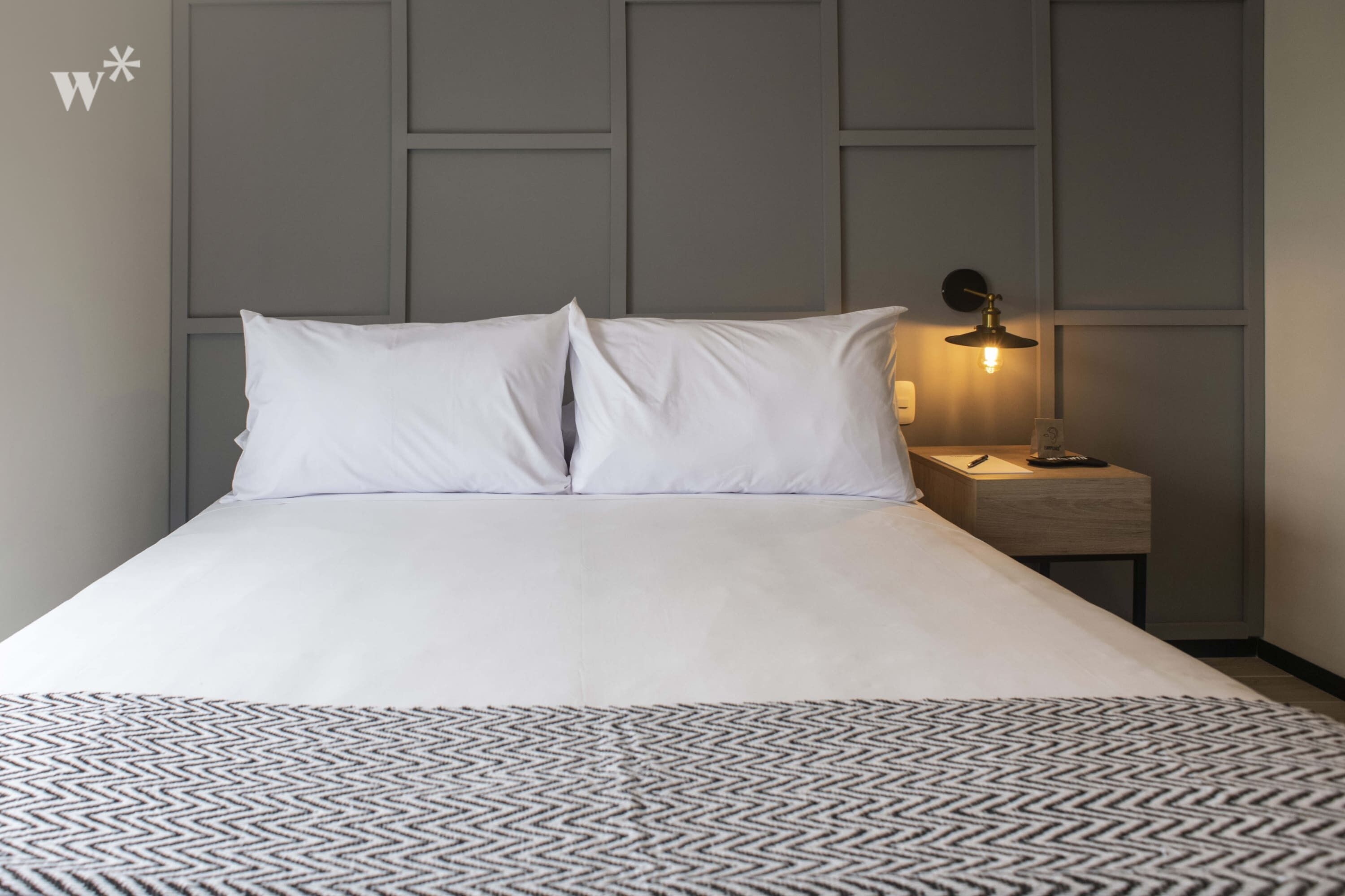 Una cama bien tendida con sábanas blancas y un plaid estampado, una mesita de noche con lámpara, y una pared con diseño de panel geométrico.