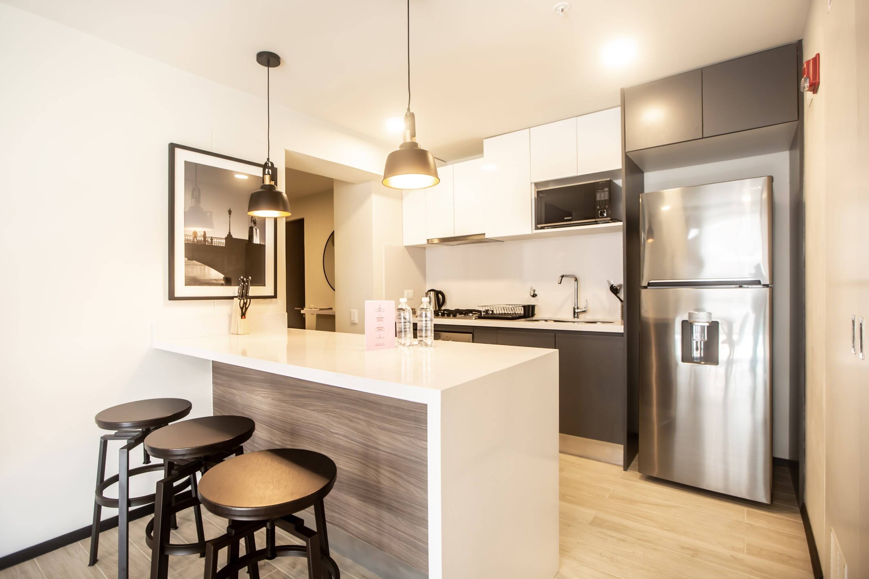 Una cocina moderna con una isla central, taburetes de bar, electrodomésticos de acero inoxidable, iluminación colgante y un cuadro en la pared.