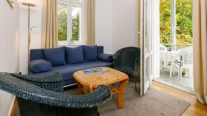 A cozy living room with a blue sofa, a wicker chair, and a wooden coffee table. There is a large window with curtains, and a balcony with outdoor furniture is visible. - App.11 Zinnowitzer Hof by Interhome (Zinnowitz)