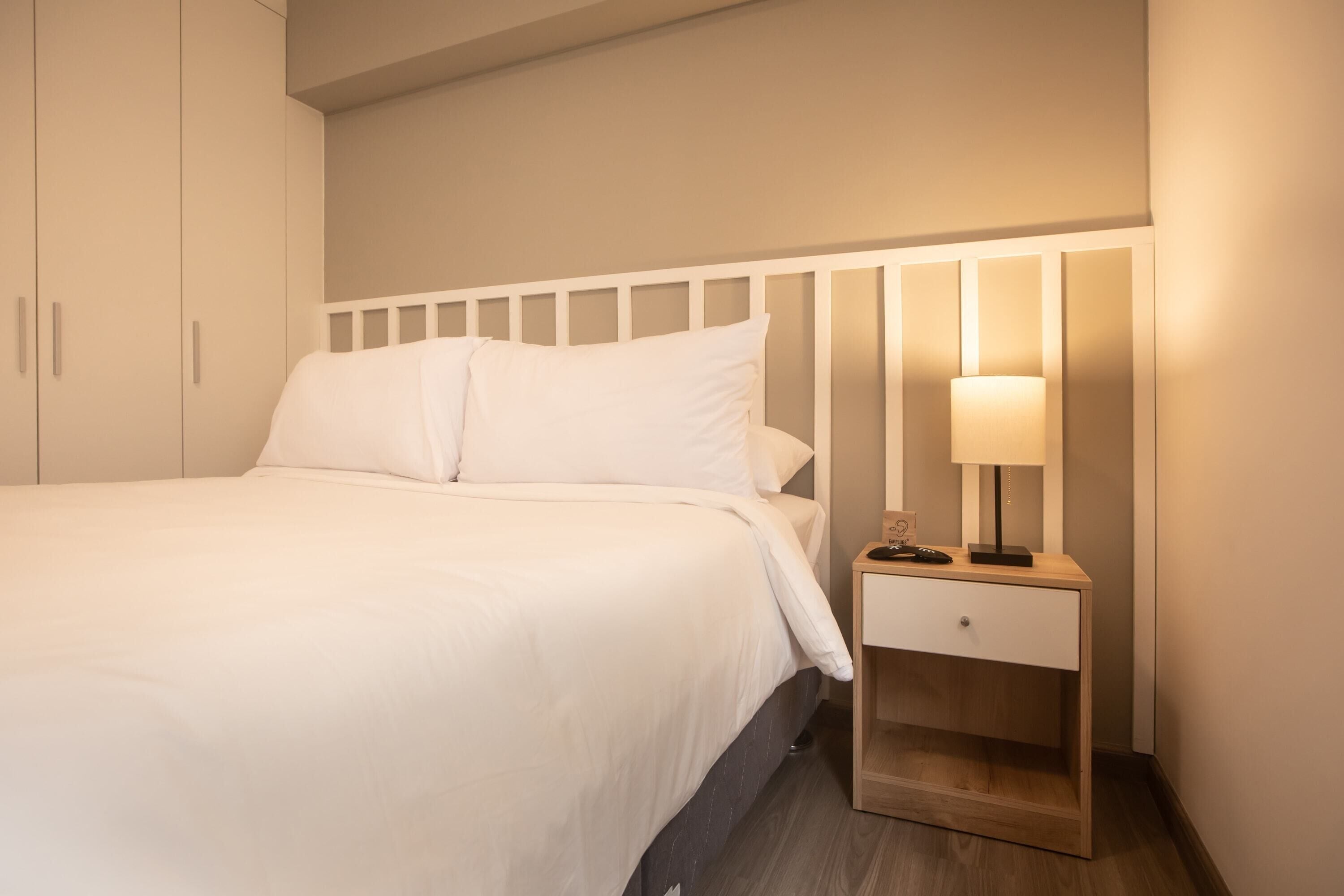 Una cama bien hecha con sábanas blancas, una mesita de noche con una lámpara y una mesita auxiliar de madera.