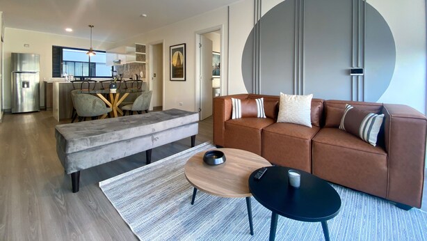Una sala de estar moderna con un sofá de cuero marrón, un banco gris y una mesa de centro redonda de madera.