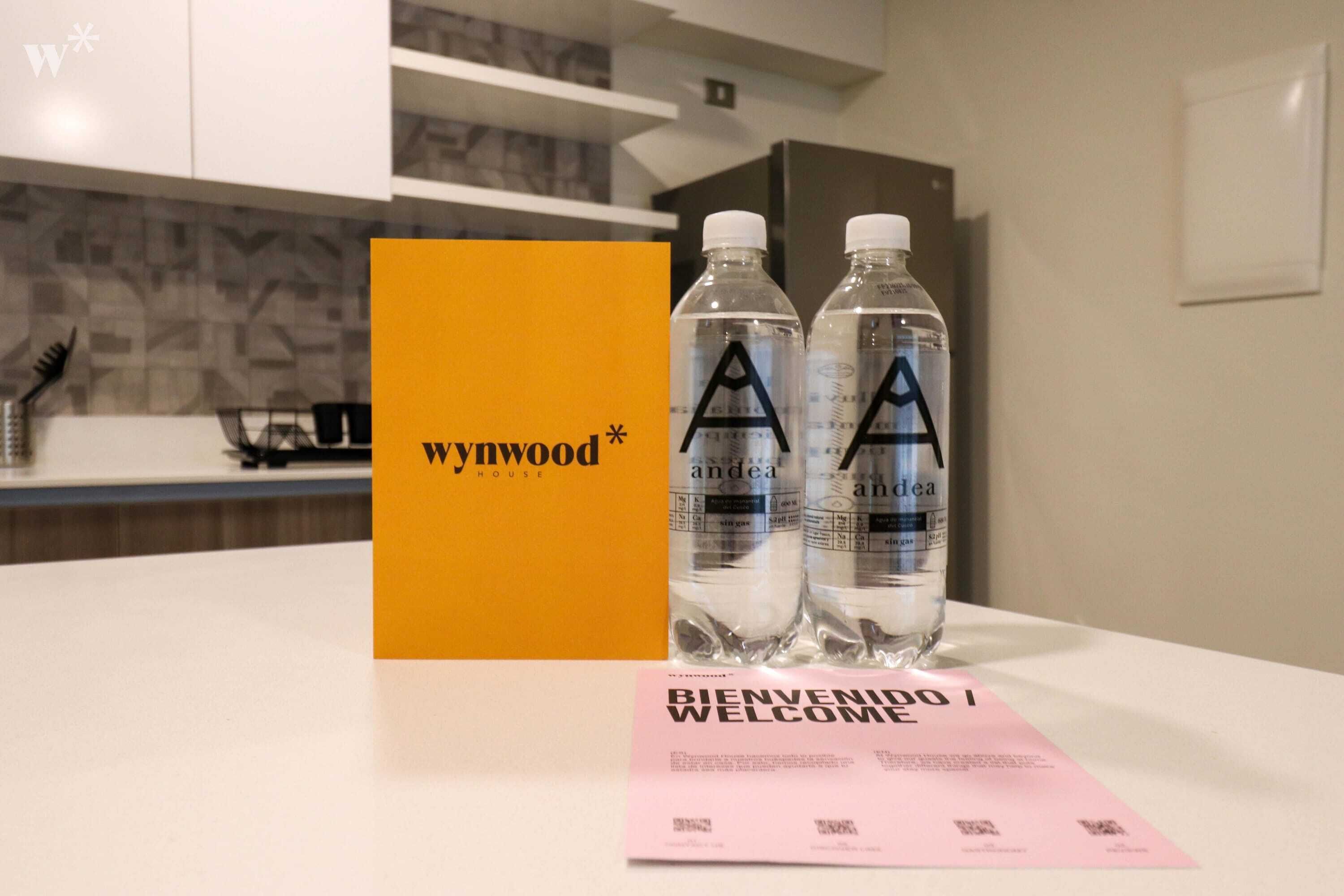 A hotel room with a yellow card labeled "Wywood House" and two water bottles with the "A" logo. A pink welcome card is also visible.