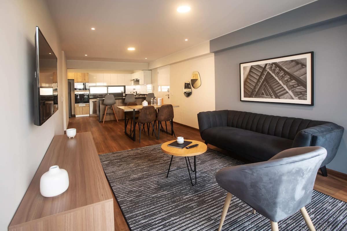 Ein modernes Wohnzimmer mit einer schwarzen Couch, einem runden Couchtisch und einem grauen Sessel. Ein Flachbildfernseher ist an der Wand montiert, und ein gerahmtes Bild hängt über der Couch.