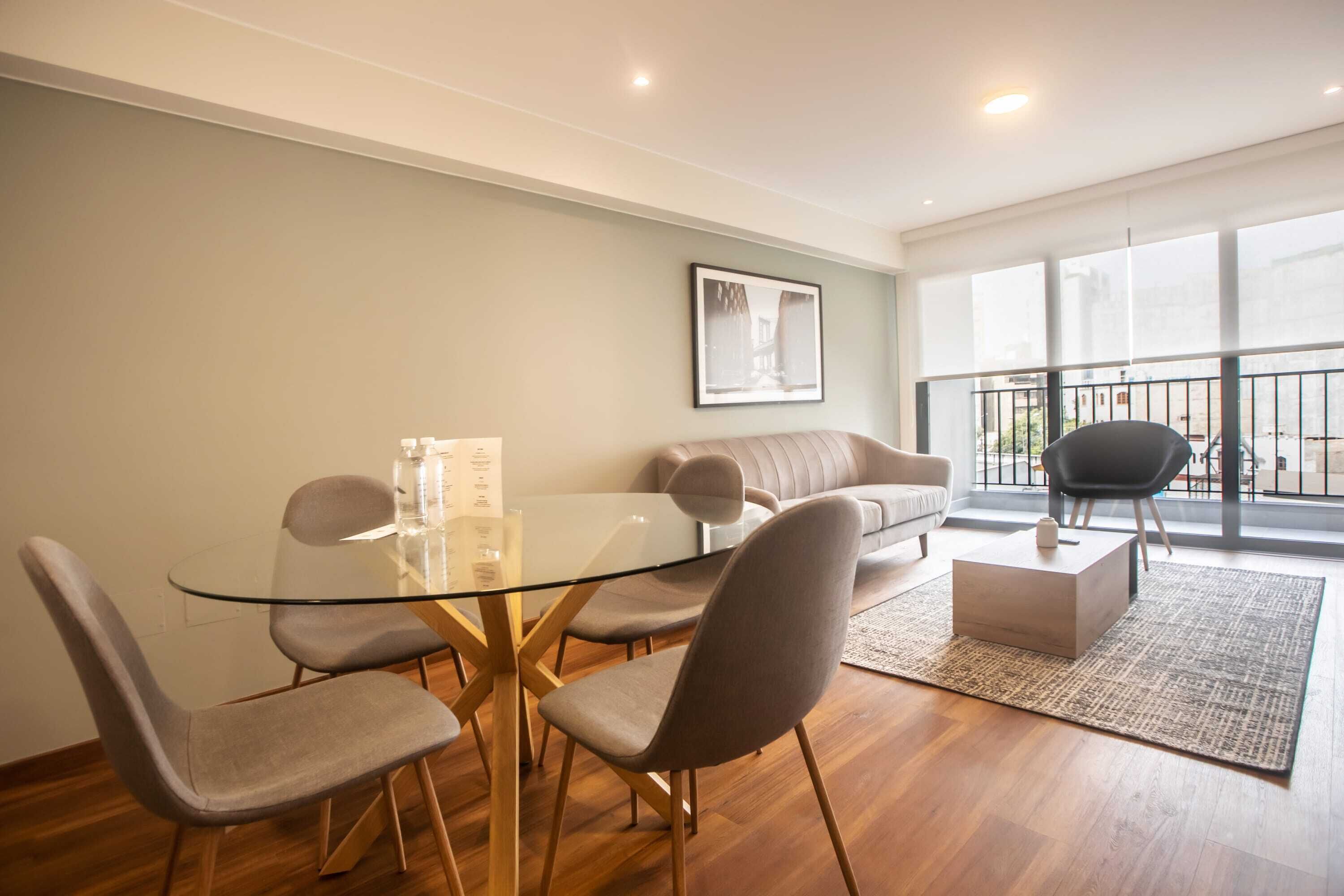 A modern living room with a glass-top dining table, grey upholstered chairs, a grey sofa, and a black armchair.