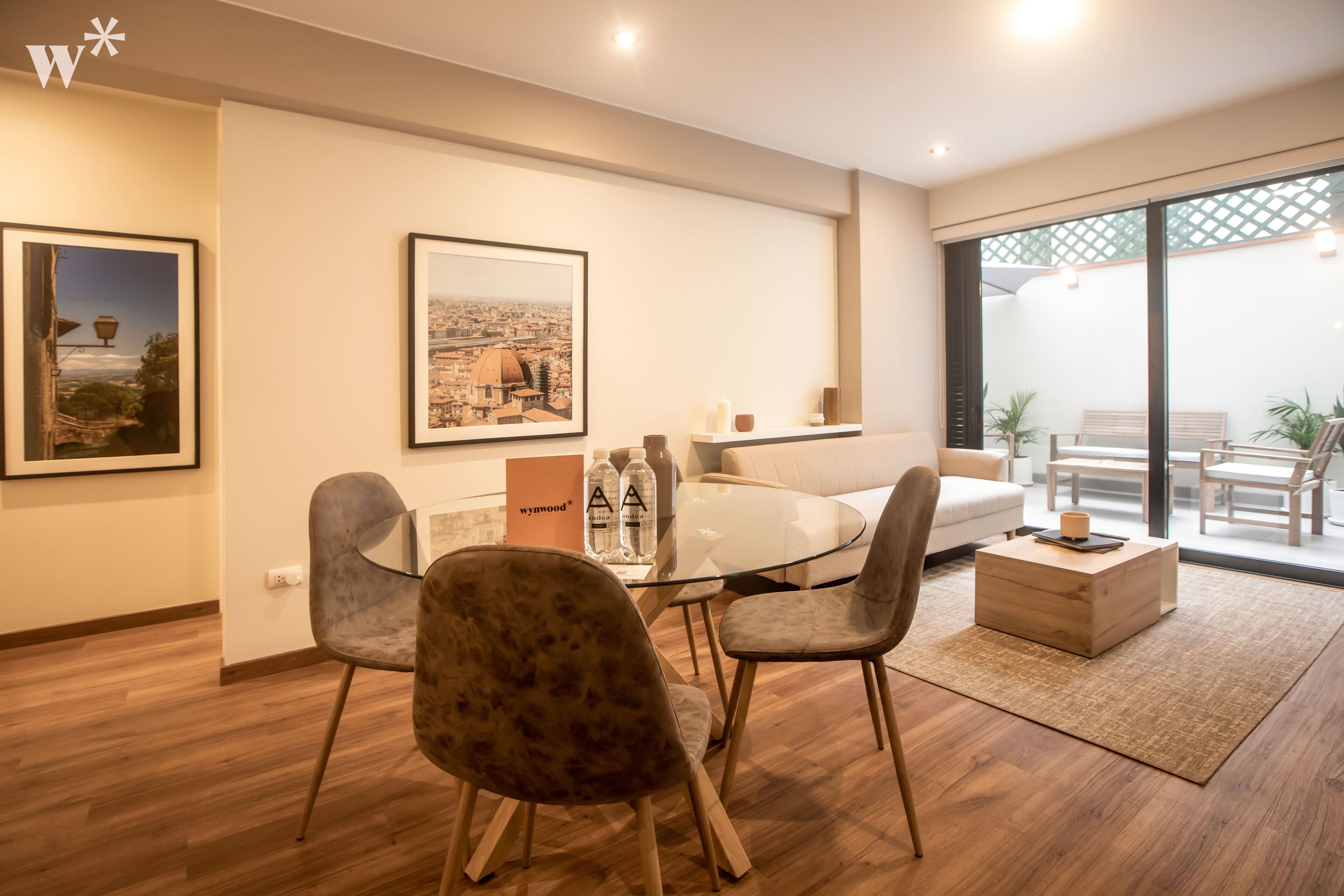 A modern living room with a glass dining table, comfortable seating, and a large window offering a view of the outdoors.
