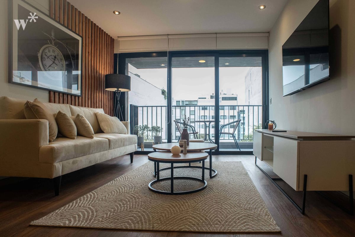 Ein modernes Wohnzimmer mit einer Couch, einem Couchtisch, einem Teppich und einem großen Fenster mit Blick auf Gebäude.