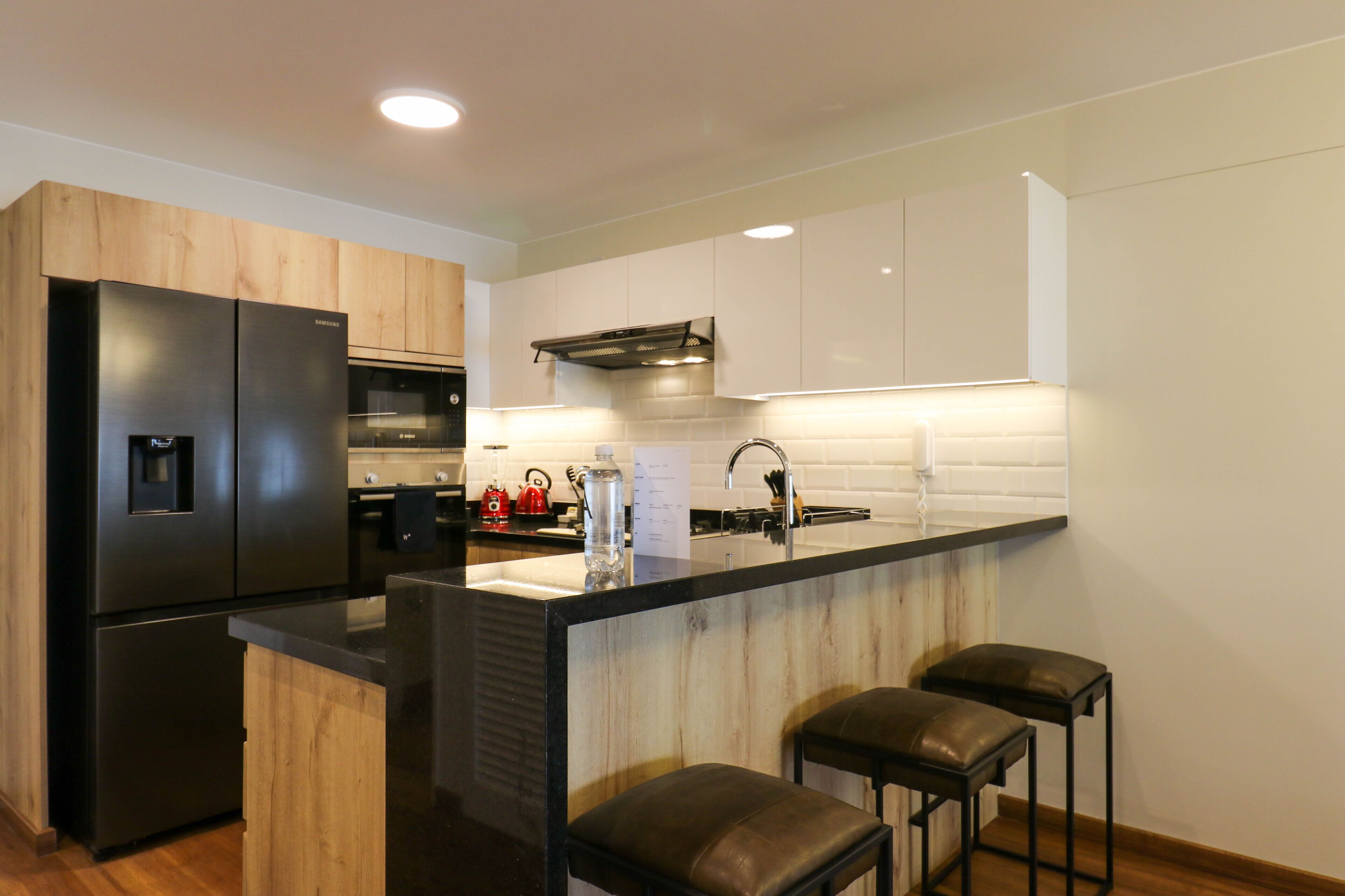 A modern kitchen with a black refrigerator, wooden cabinets, a black countertop, and three black bar stools.