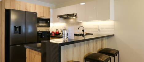 A modern kitchen with a black refrigerator, wooden cabinets, a black countertop, and three black bar stools.