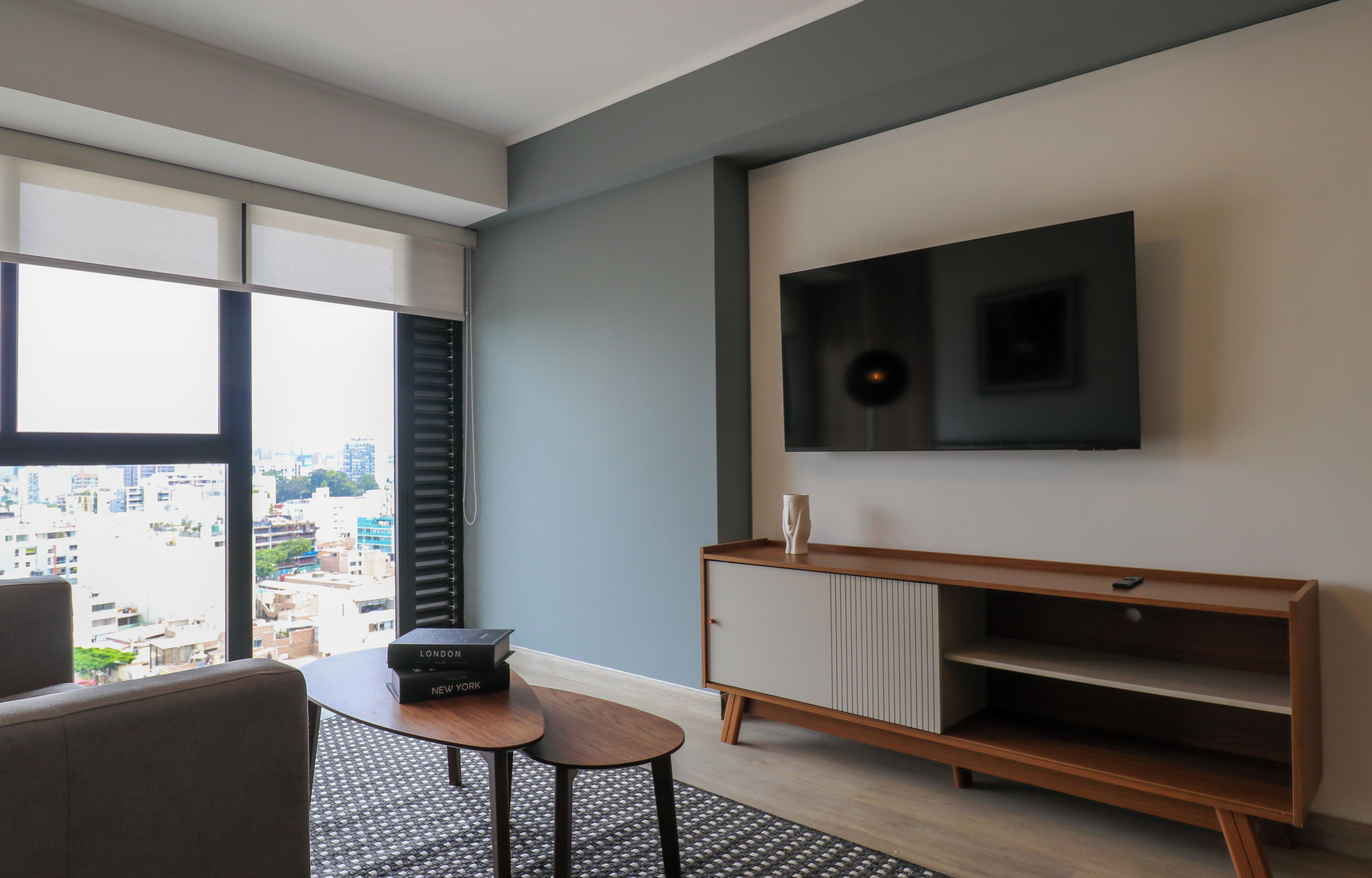 A modern living room with a large window, a flat-screen TV mounted on the wall, a wooden entertainment unit, and a small table with a remote control.