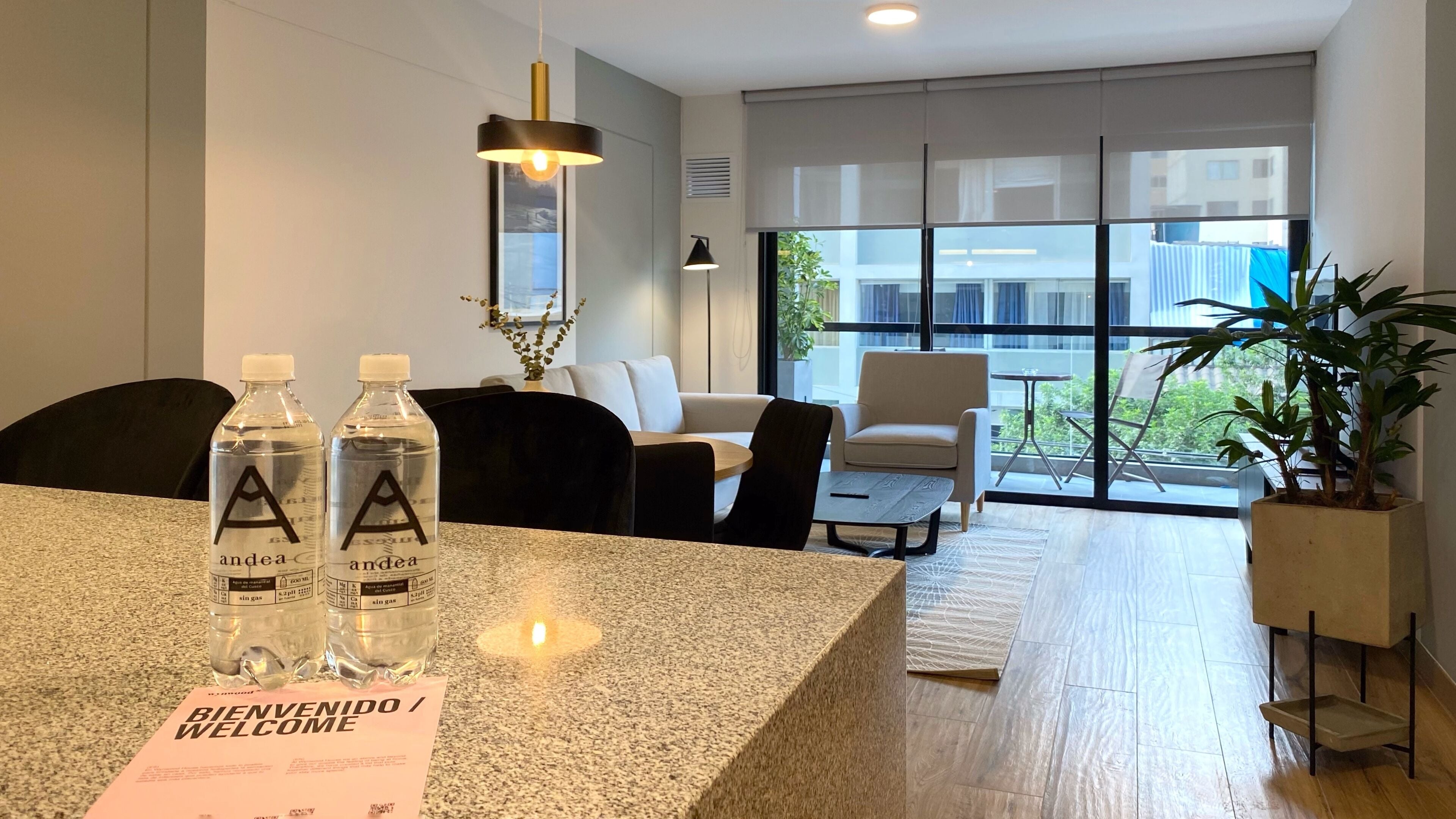 Een moderne hotel lobby met een receptiebalie van graniet, twee waterflessen, een welkomstbord, een bank, een bijzettafeltje en een groot raam met uitzicht op de stad.