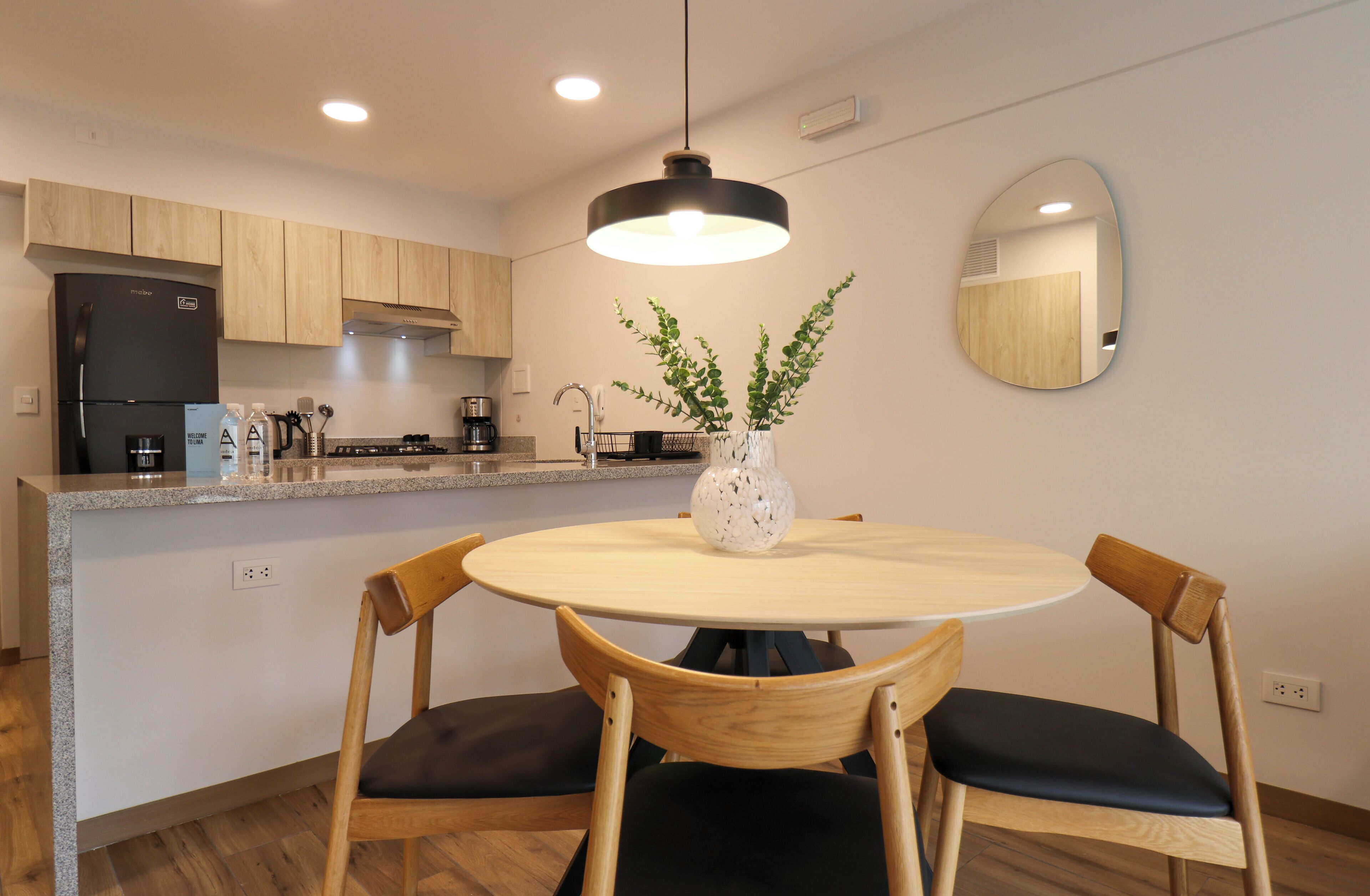 Une cuisine moderne avec des meubles en bois, un réfrigérateur noir, une table de salle à manger ronde avec deux chaises, un luminaire suspendu et un miroir au mur.