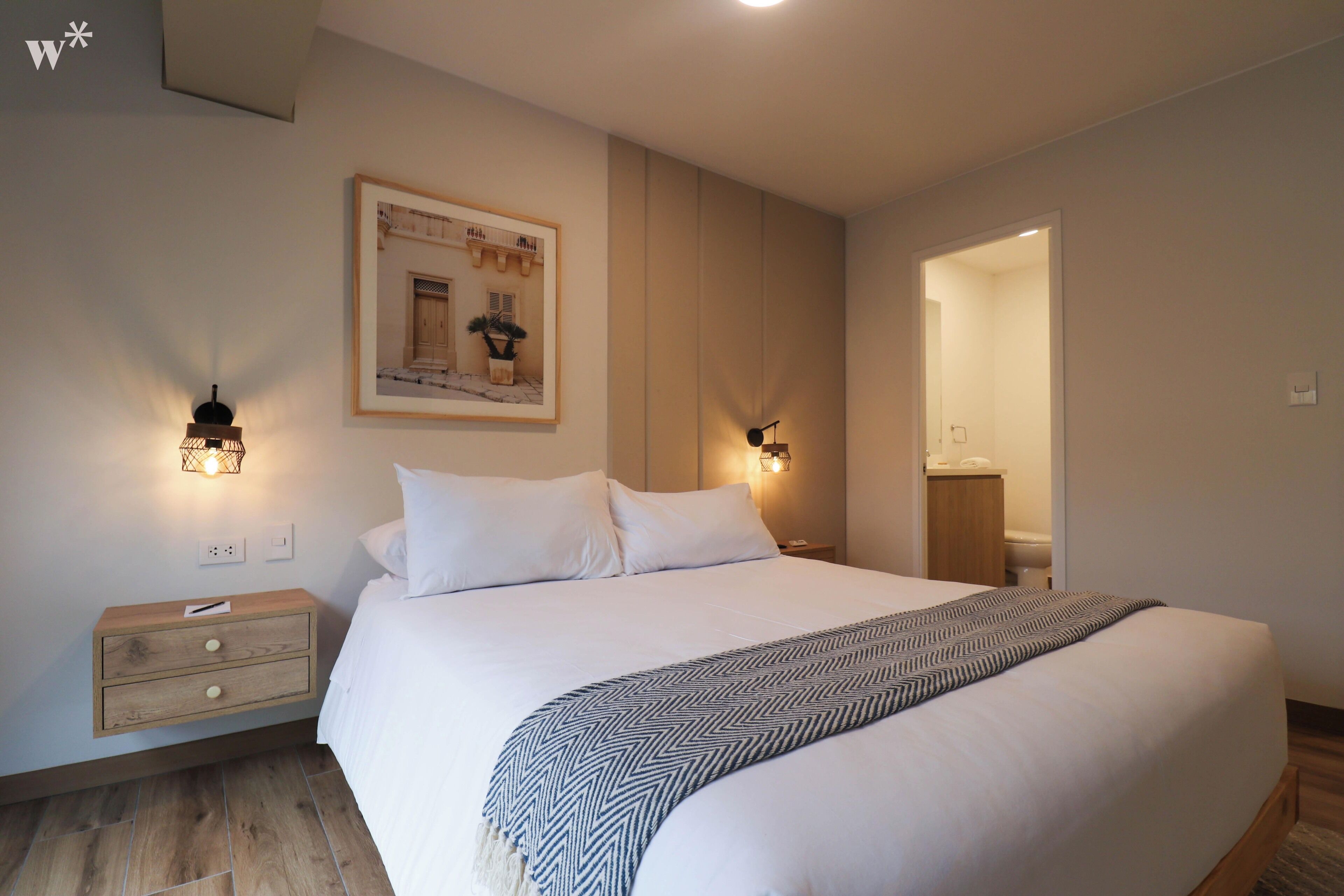A neatly made bed with a patterned throw, bedside lamps, a framed picture on the wall, and a wooden nightstand.