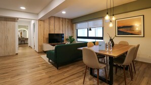 A modern living room with a wooden floor, a green sofa, a dining table with chairs, a TV, and a large window with a view of a sunset. - Wynwood Colon in Miraflores (Lima)