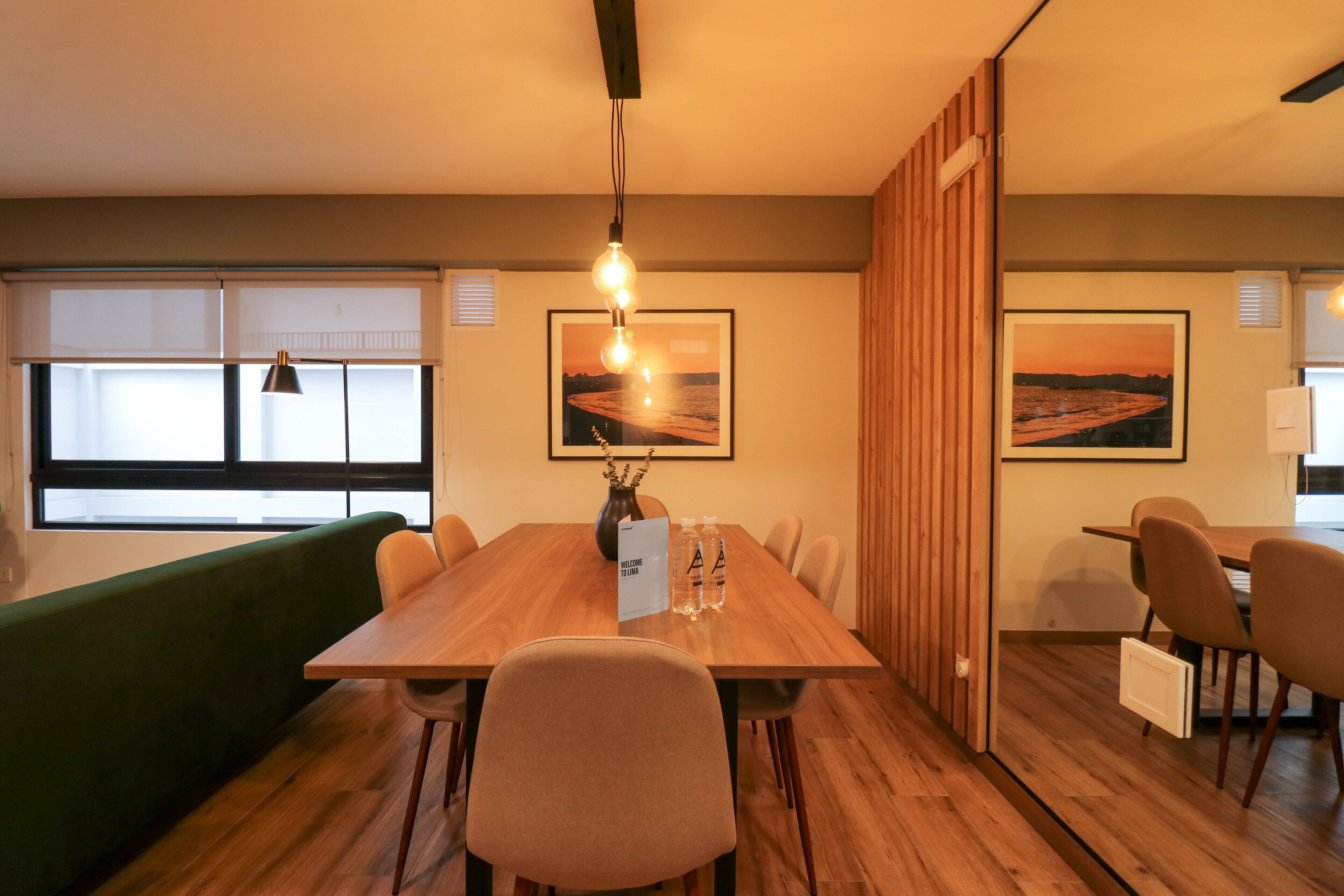 A modern dining area with a wooden table, chairs, and a green couch. There are two framed pictures on the wall, a vase with branches, and a welcome sign.