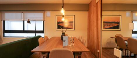 A modern dining area with a wooden table, chairs, and a green couch. There are two framed pictures on the wall, a vase with branches, and a welcome sign.