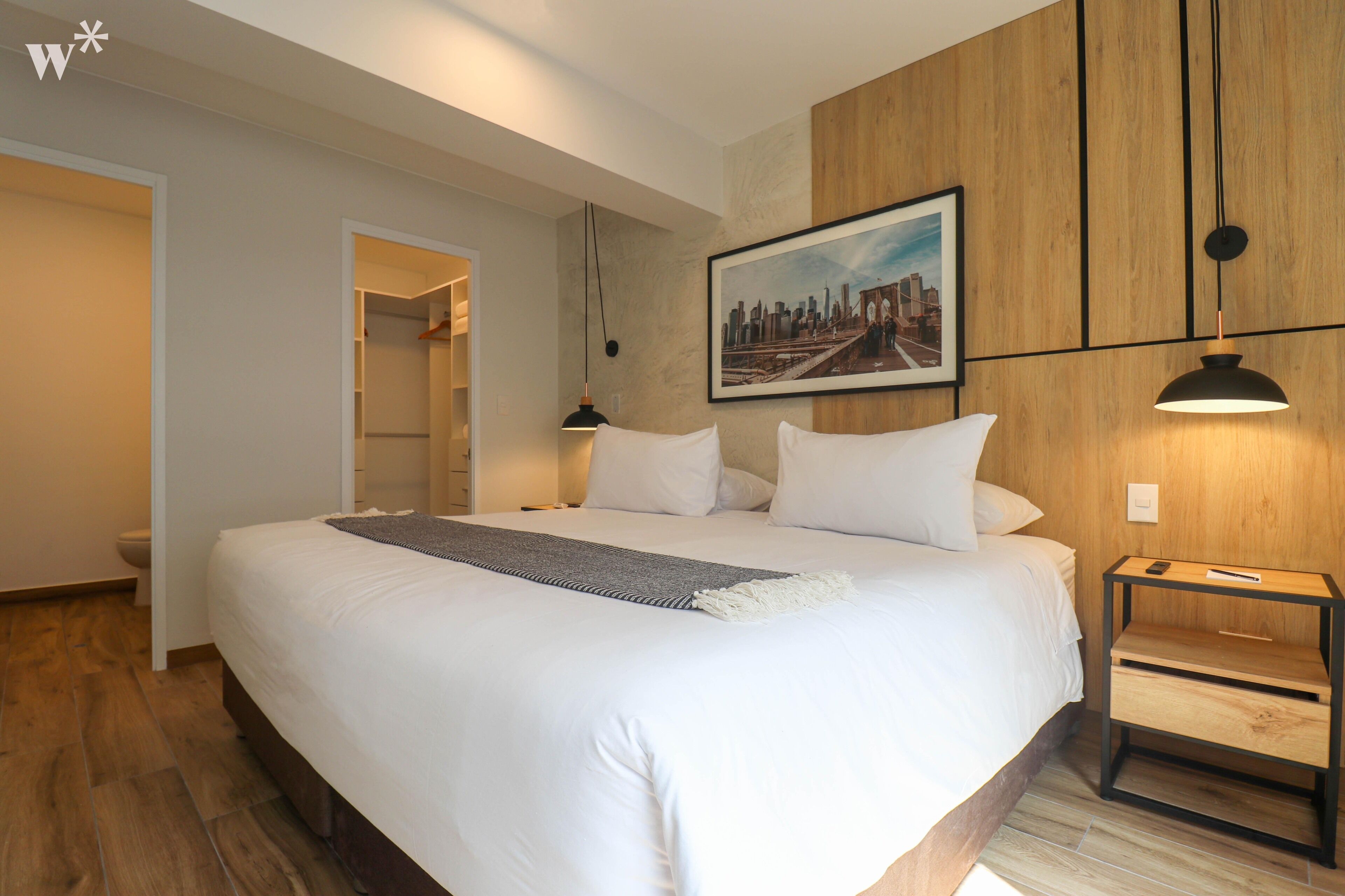 A hotel room with a large bed, a wooden panel wall, a framed cityscape picture, a bedside table with a lamp, and a walk-in closet.