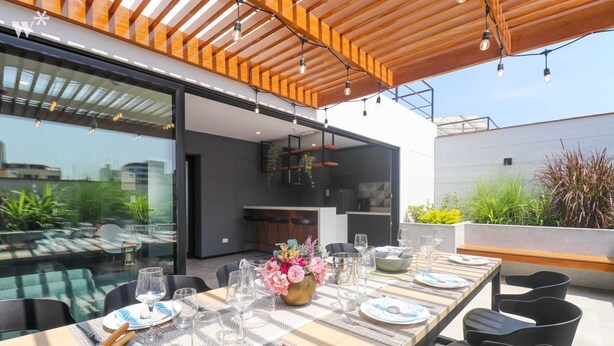An outdoor dining area with a long table set for six, surrounded by black chairs. There is a wooden pergola overhead with string lights, and a modern building with large windows and a glass wall is visible in the background. - Wynwood Colon in Miraflores (Lima)