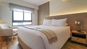 A modern bedroom with a large bed, a bedside table with a lamp, a chair, and a window with blinds. - Wynwood Colon in Miraflores (Lima)