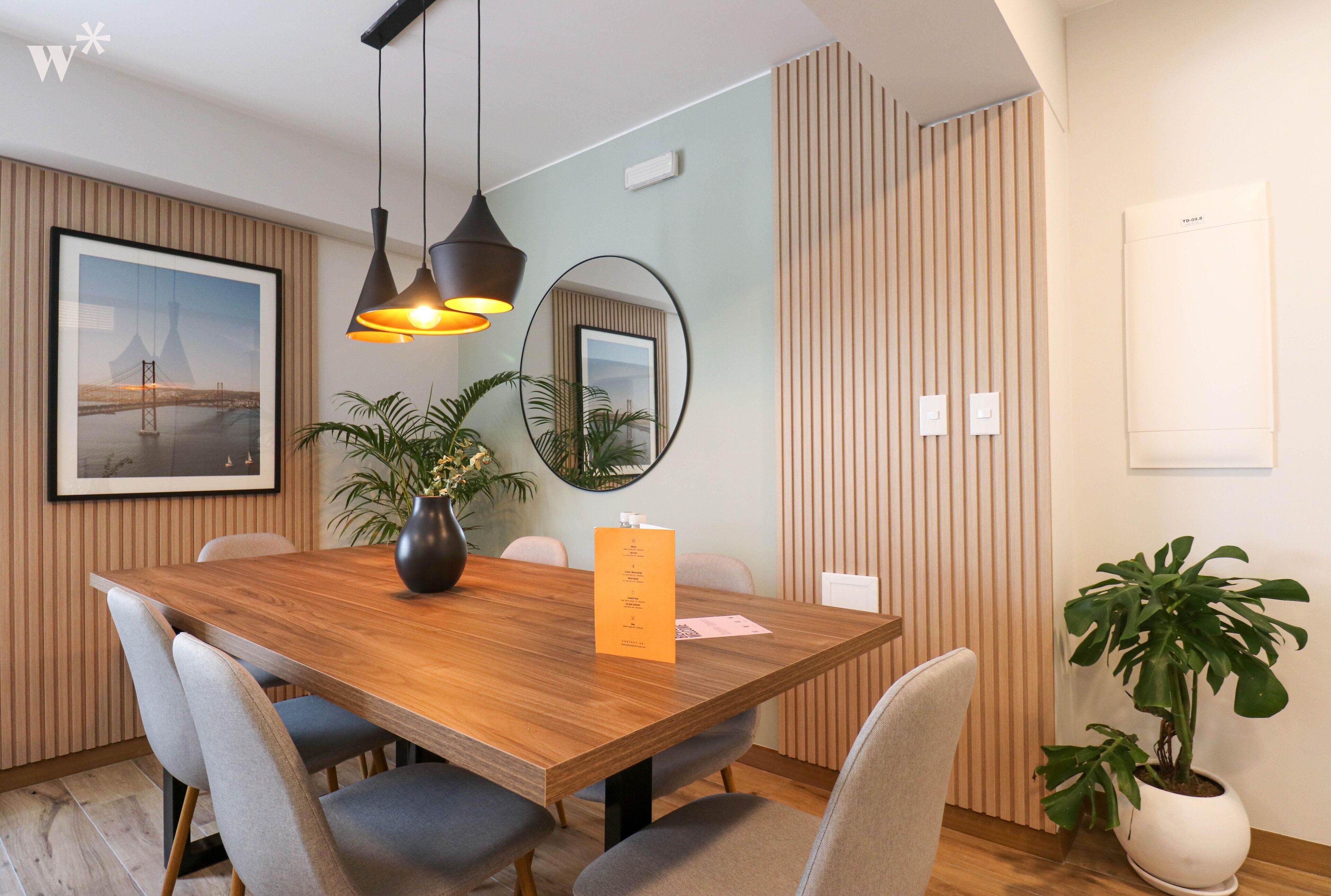 Un comedor moderno con una mesa de madera, sillas grises, iluminación colgante y una planta en maceta.