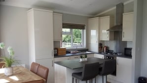 A modern kitchen with a dining area, featuring a large island, bar stools, and a view of the outdoors. - Cottage in Brookland near Romney Marsh Beach (Fairfield)