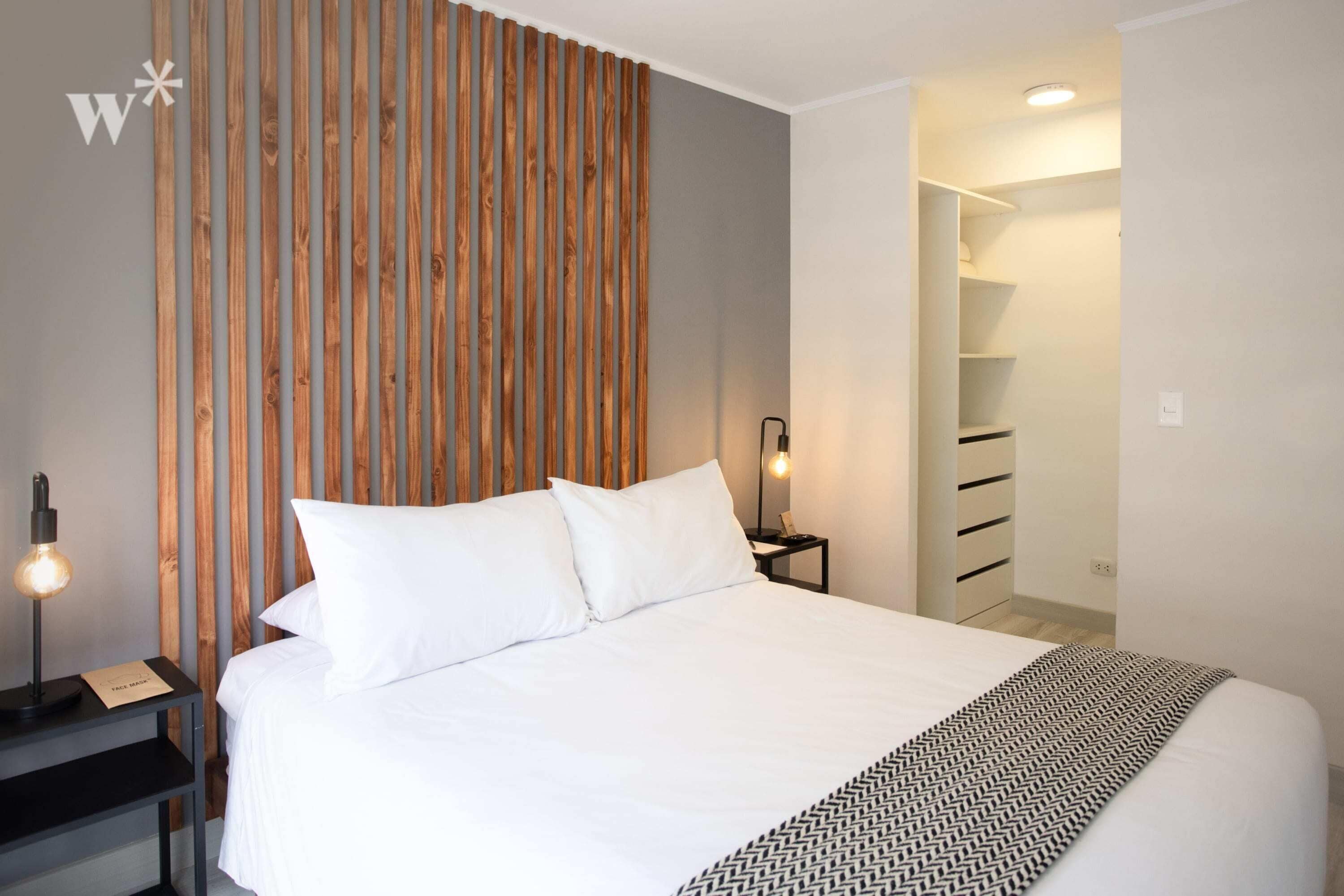 Habitación de hotel moderna con una cama grande, cabecera de listones de madera, mesita de noche con lámpara y armario empotrado.