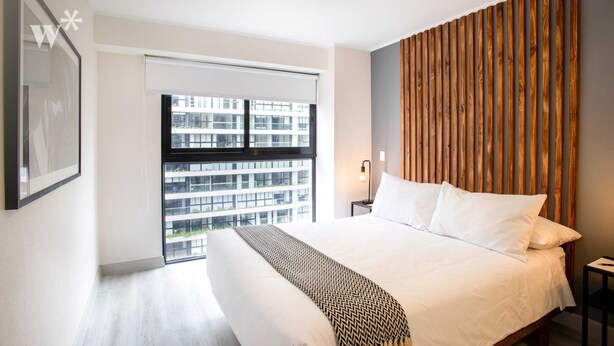 A modern bedroom with a large bed, a wooden headboard, a view of a building through a window, and a framed picture on the wall. - Wynwood Dean Valdivia (Lima)