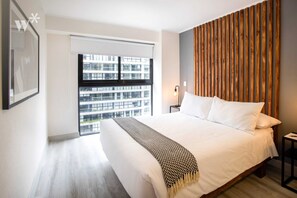 A modern bedroom with a large bed, a wooden headboard, a view of a building through a window, and a framed picture on the wall.