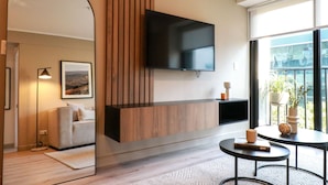 A modern living room with a flat-screen TV mounted on a wooden panel, a sofa, a coffee table, and a large window offering a view of the outdoors. - Wynwood Dean Valdivia (Lima)