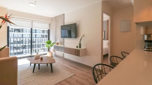 A modern living room with a large window, a flat-screen TV mounted on the wall, a dining area with a wooden table, and a kitchen area with a coffee machine. - Wynwood Dean Valdivia (Lima)