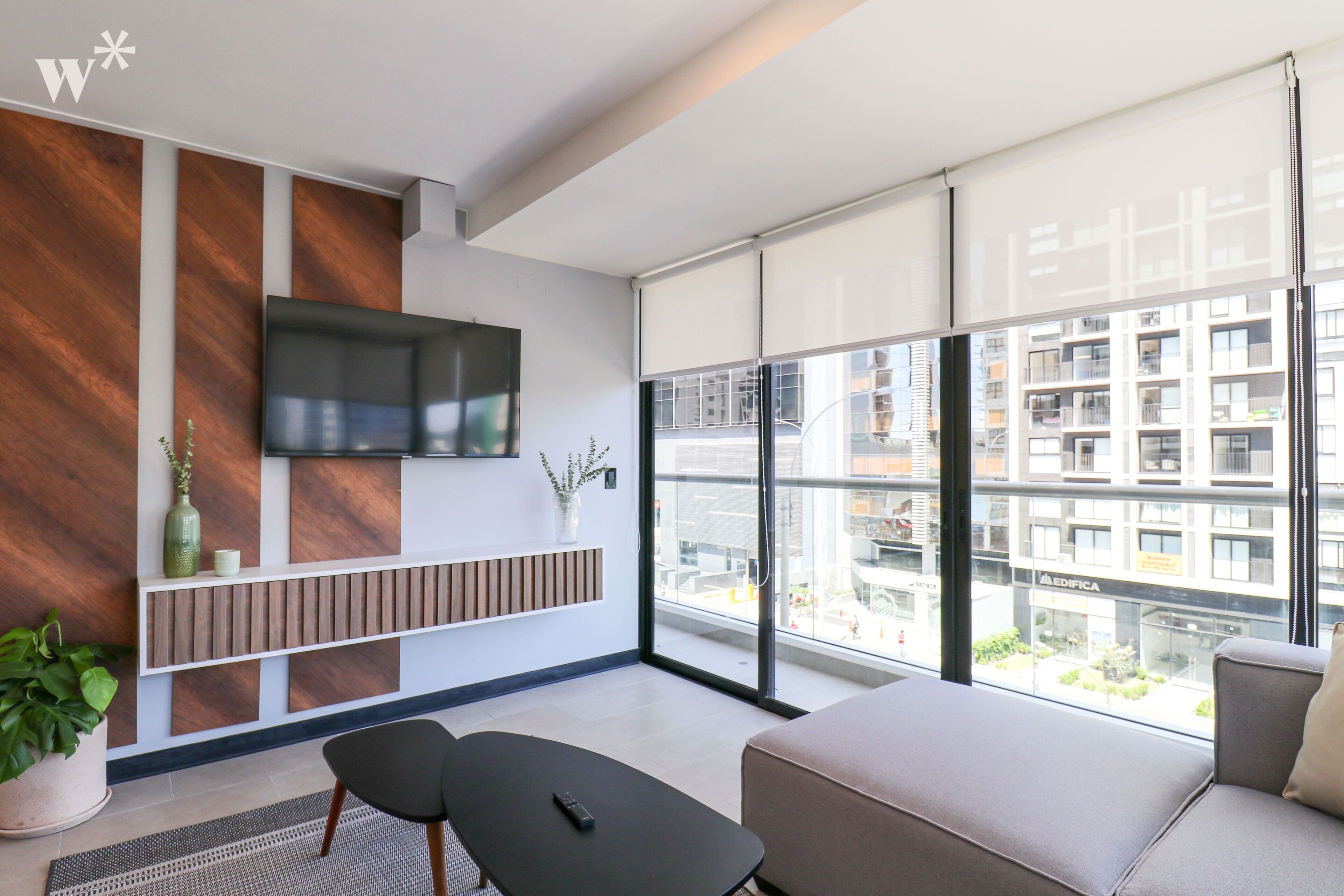 Ein modernes Wohnzimmer mit einer Couch, einem an der Wand montierten Fernseher, einer Topfpflanze und Blick auf eine Stadtlandschaft durch große Fenster.