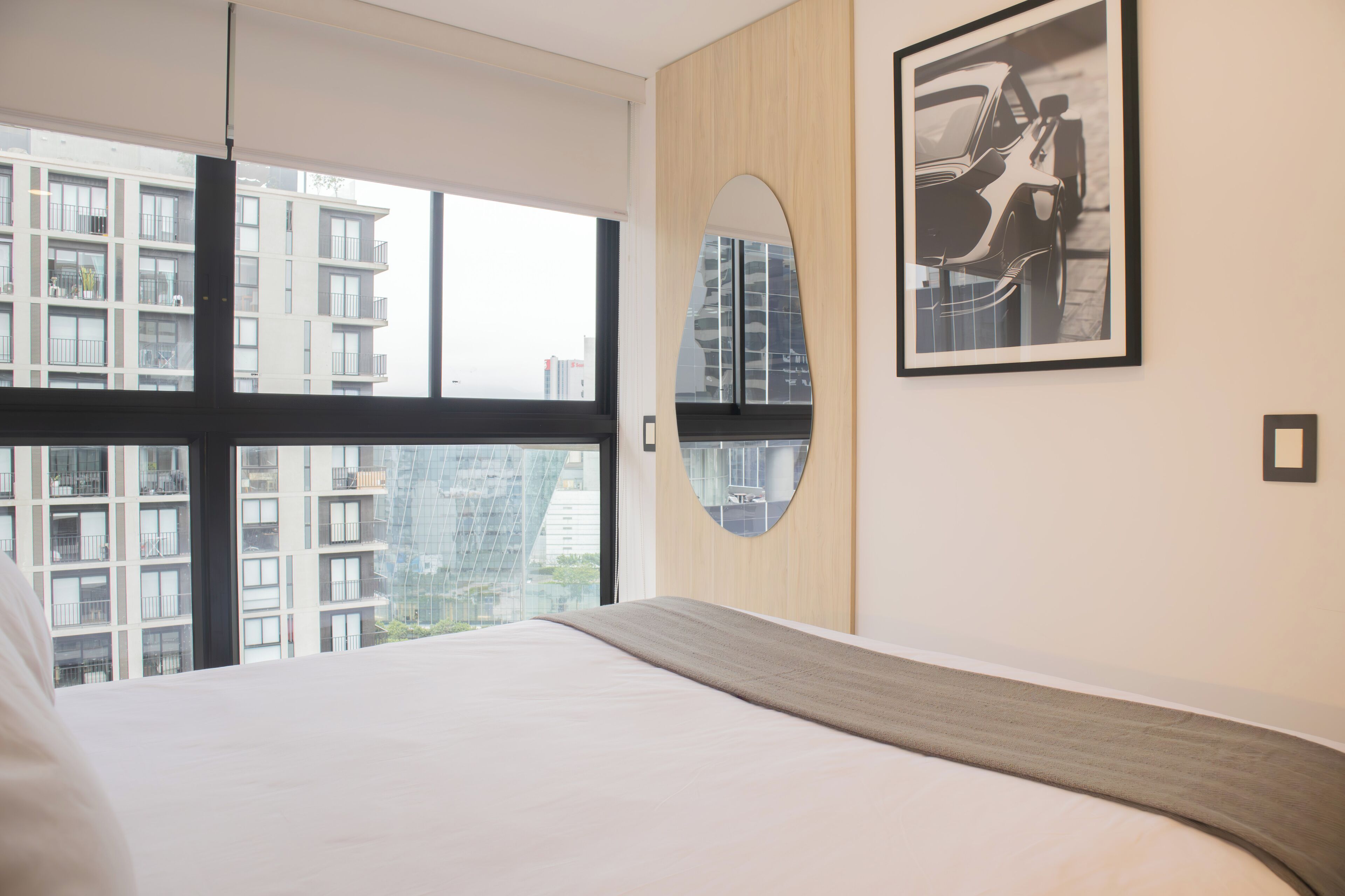 Une chambre d’hôtel moderne avec un grand lit, une vue sur les gratte-ciel, un miroir rond et un tableau encadré au mur.