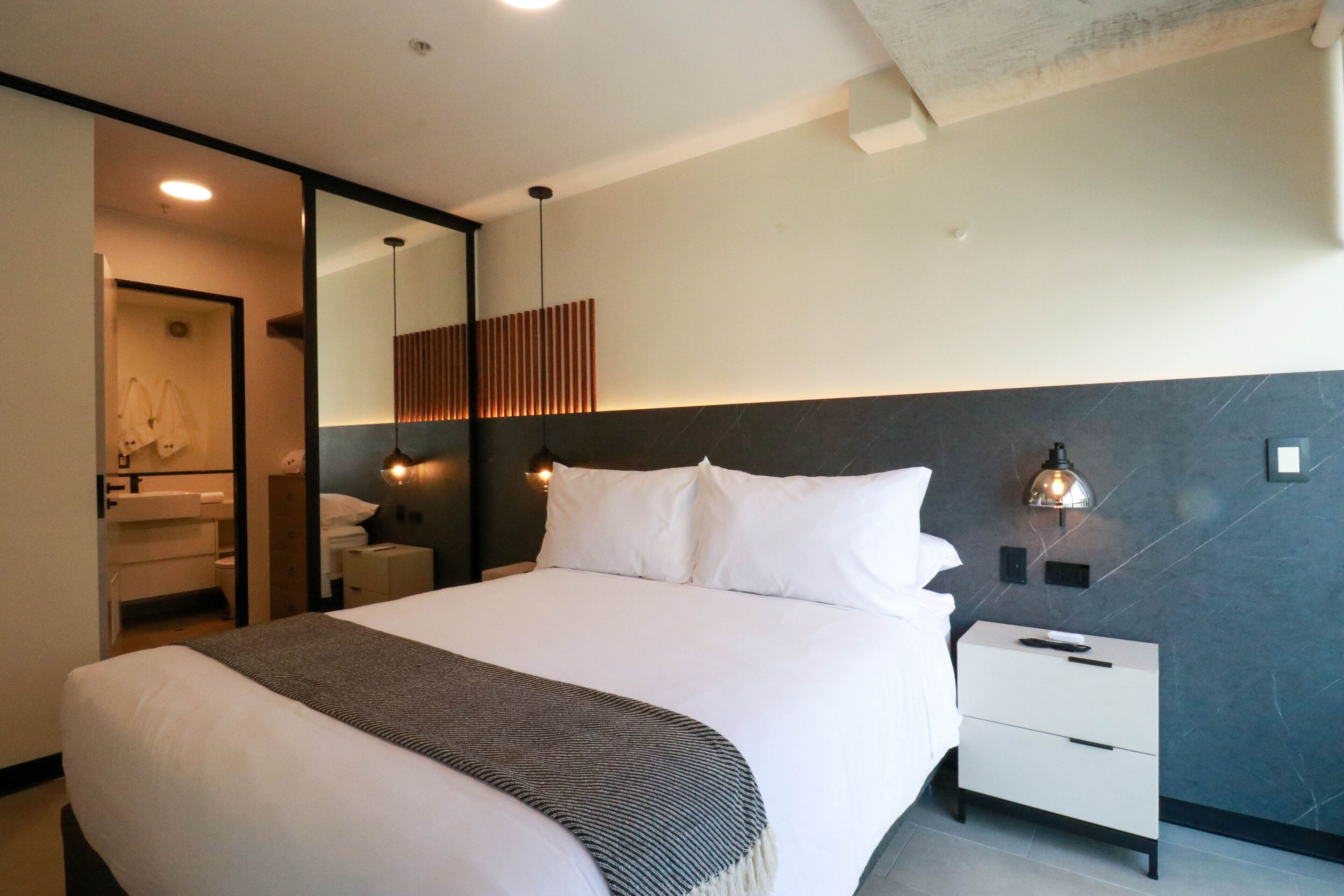 A modern hotel room with a large bed, a bedside table, a nightstand, a mirror, and a wall-mounted light fixture.
