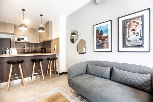 A modern kitchen with pendant lights, a bar area, and a grey sofa with cushions. - Wynwood Rivera Navarrete (Lima)