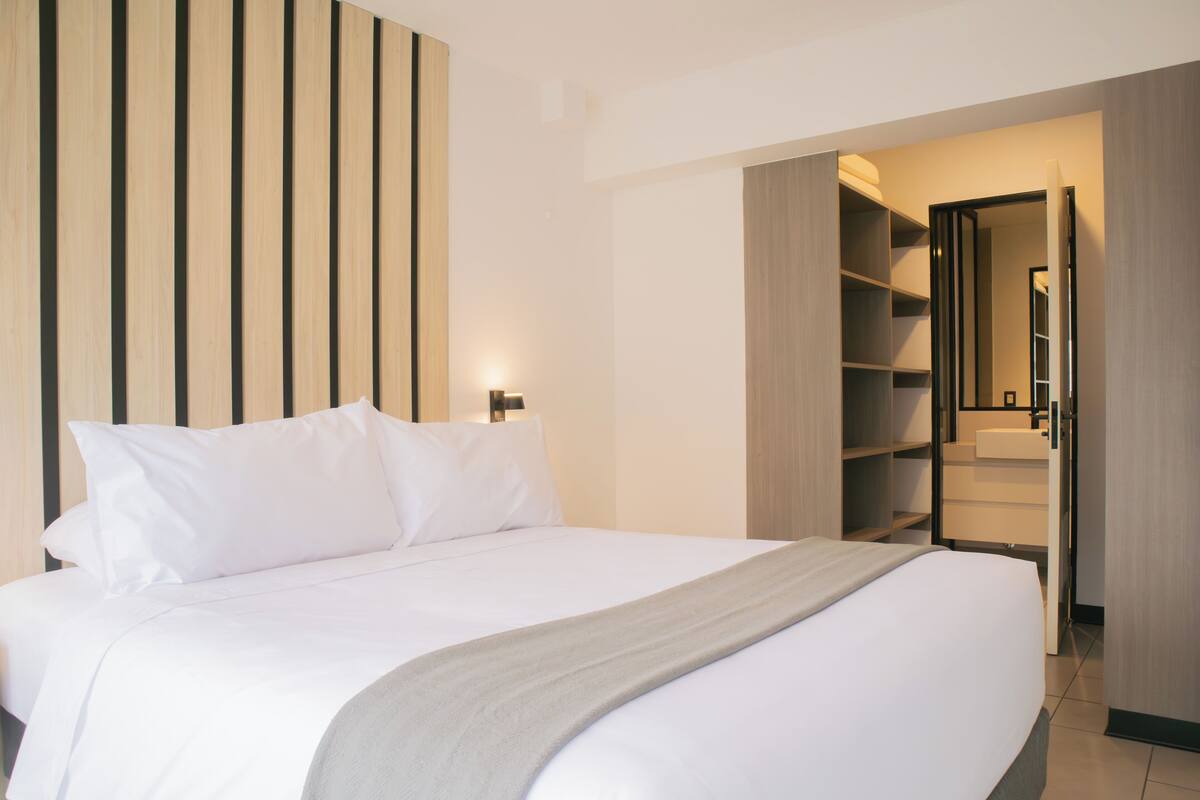 Ein ordentlich bezogenes Bett mit weißen Bettwäsche und einer grauen Decke, eine Wand aus Holzlamellen und ein begehbarer Kleiderschrank mit Regalen und einem Spiegel.