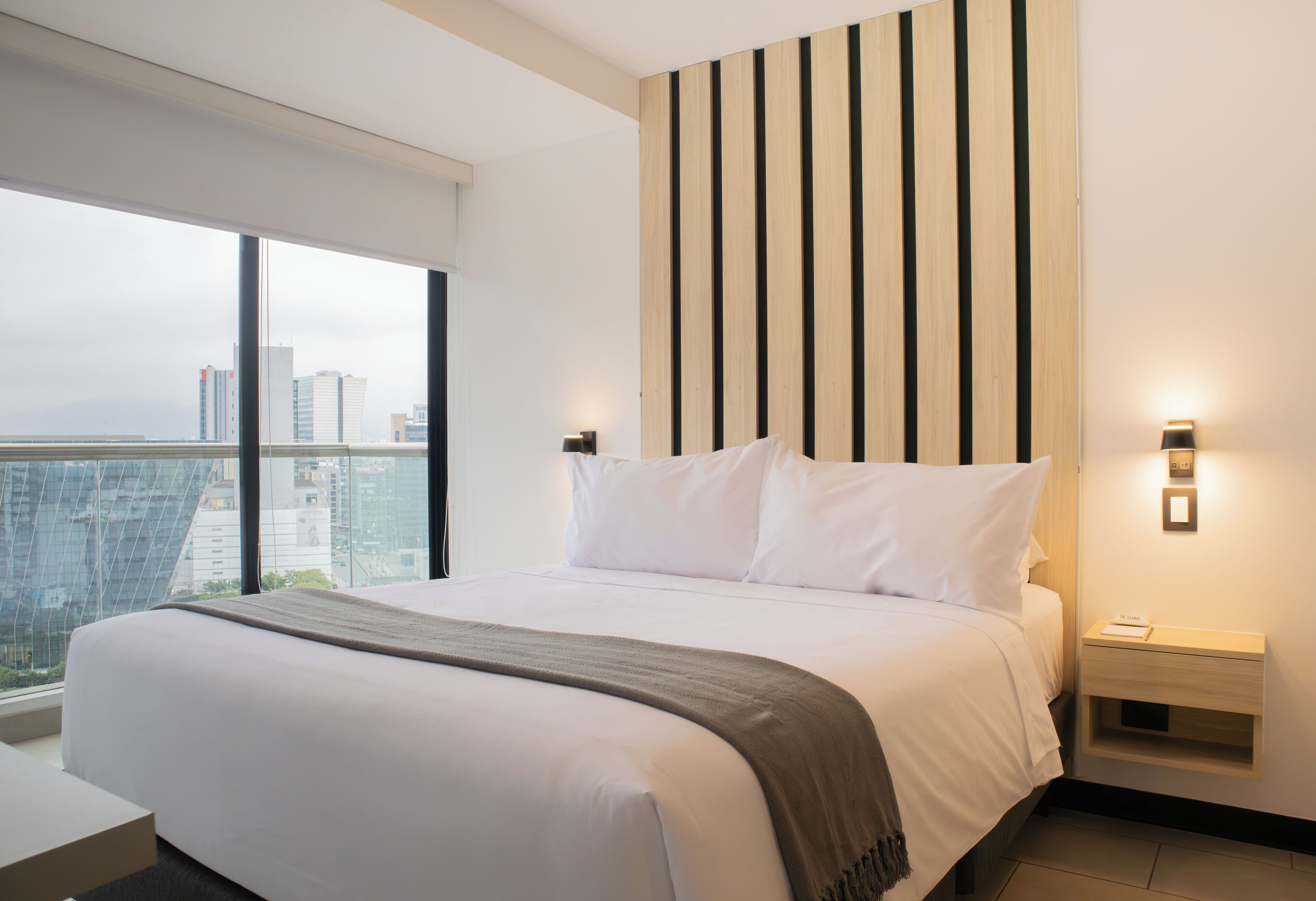 Een hotelkamer met een groot bed, nachtkastje en uitzicht op de stad door grote ramen.