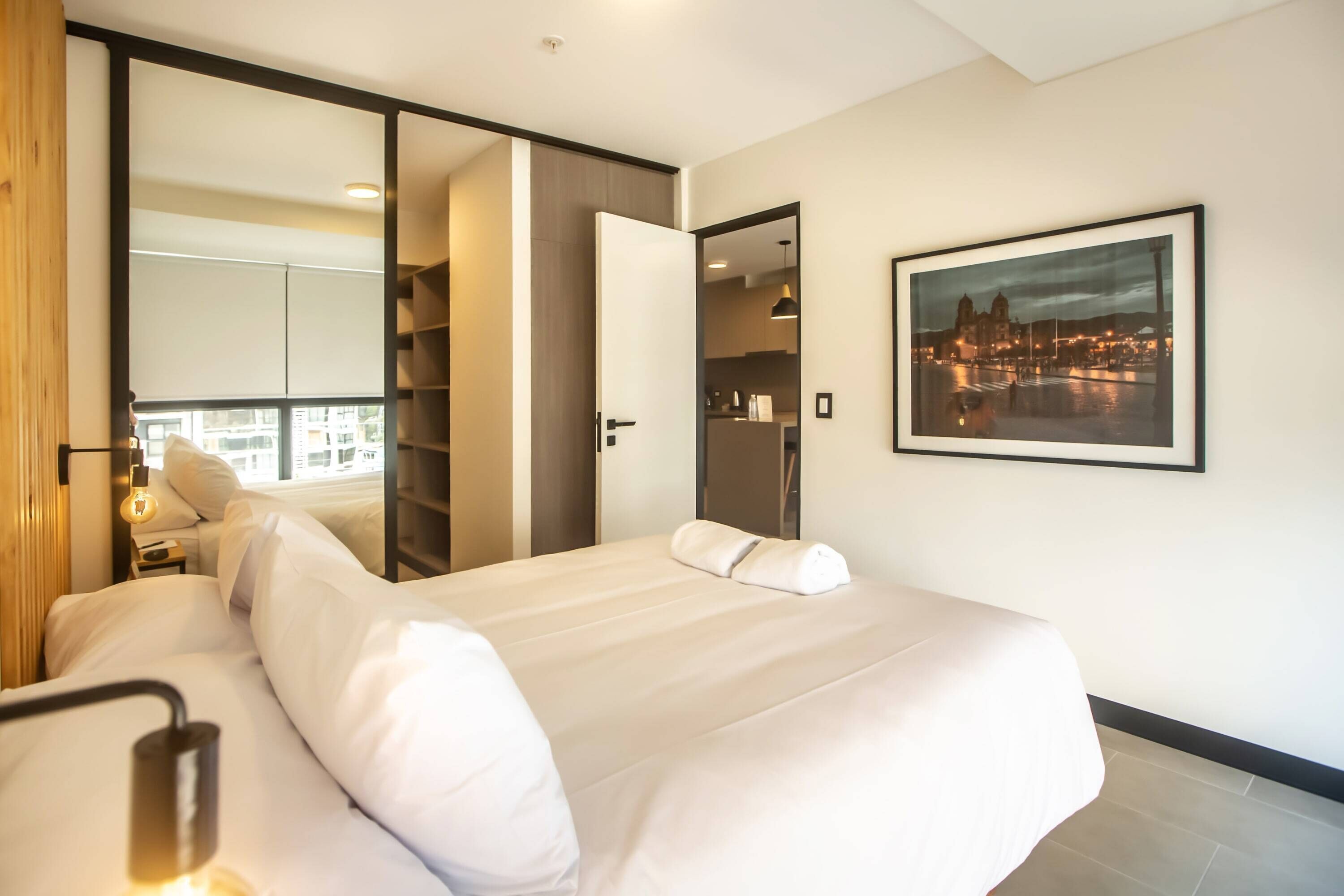A hotel room with a large bed, a wardrobe, a mirror, and a framed cityscape picture on the wall.