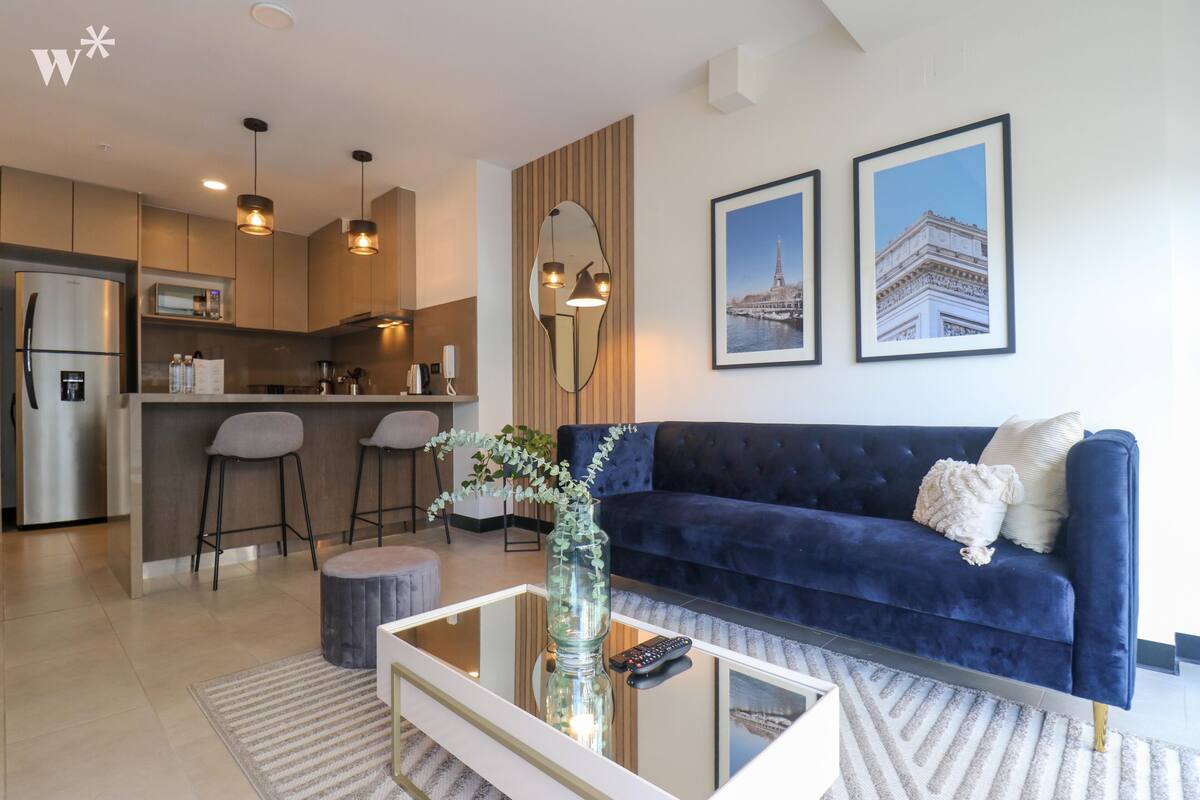 Ein modernes Wohnzimmer mit einem dunkelblauen Sofa, einem Glastisch und einer Küche im Hintergrund.