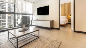 A modern hotel room with a large window, a flat-screen TV, a sofa, a bedside table, and a bed with a wooden headboard. - Wynwood Rivera Navarrete (Lima)