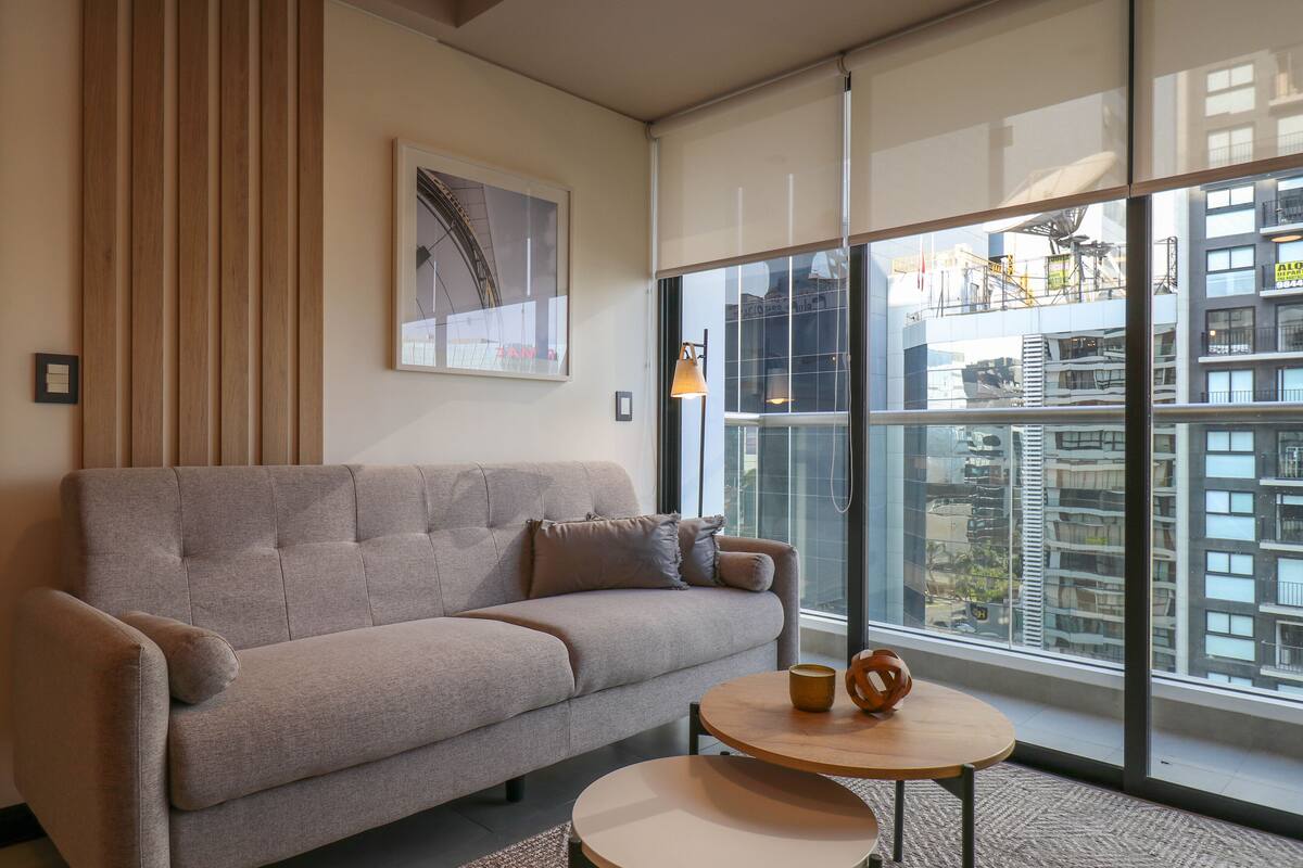 Ein modernes Wohnzimmer mit einem grauen Sofa, einem runden Holztisch und einem großen Fenster mit Blick auf eine Stadtlandschaft.