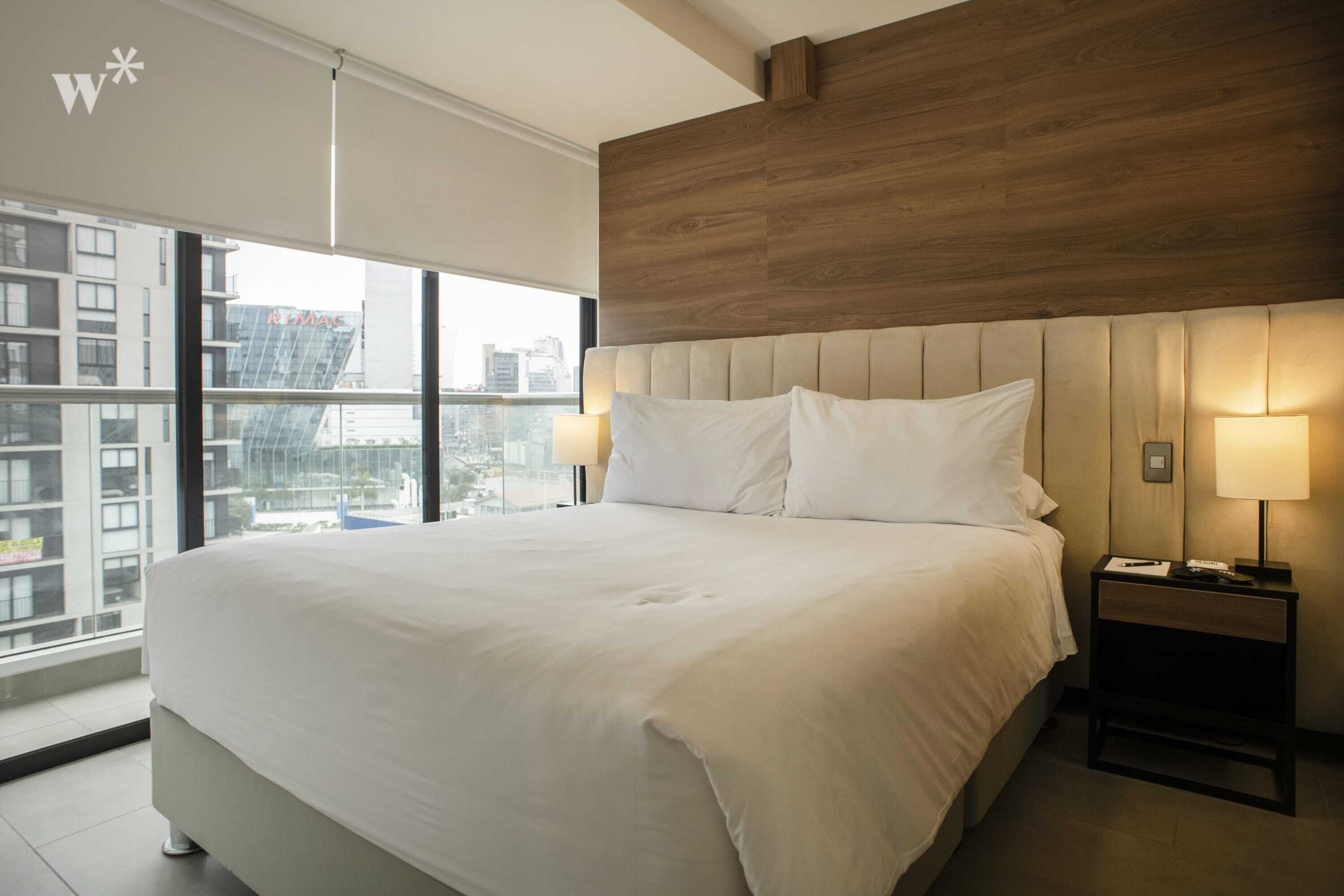 A modern hotel room with a large bed, wooden headboard, two bedside lamps, and a view of the city through large windows.