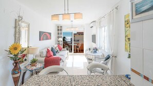 Ein helles und freundliches Wohnzimmer mit Sofa, Esstisch und Blick auf den Ozean durch eine Schiebetür.