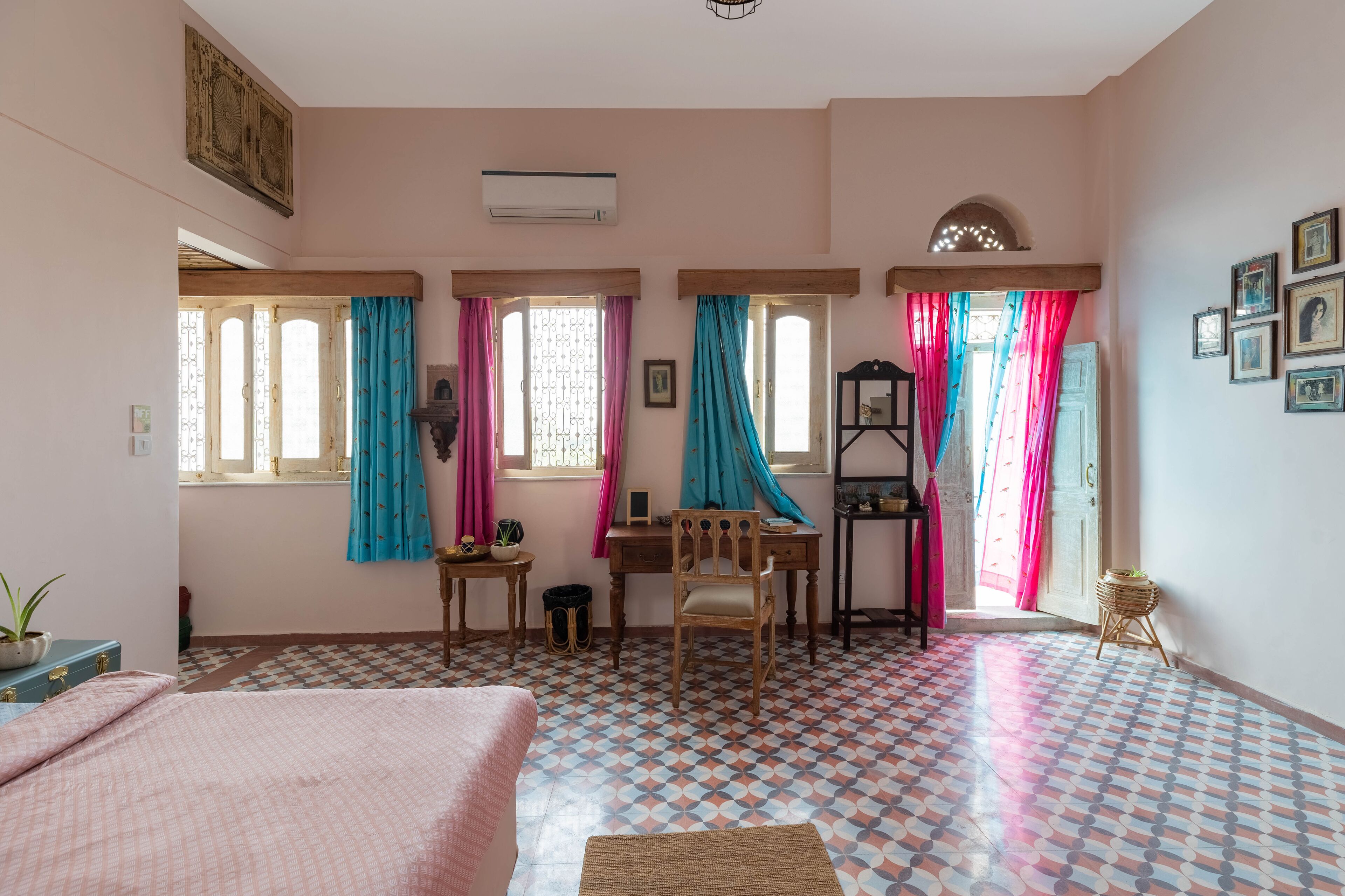 Una habitación con cama, escritorio y silla. Hay ventanas con cortinas, un radiador y una estantería en la pared.
