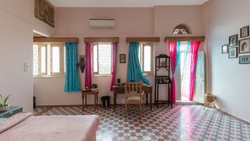 Una habitación con cama, escritorio y silla. Hay ventanas con cortinas, un radiador y una estantería en la pared.